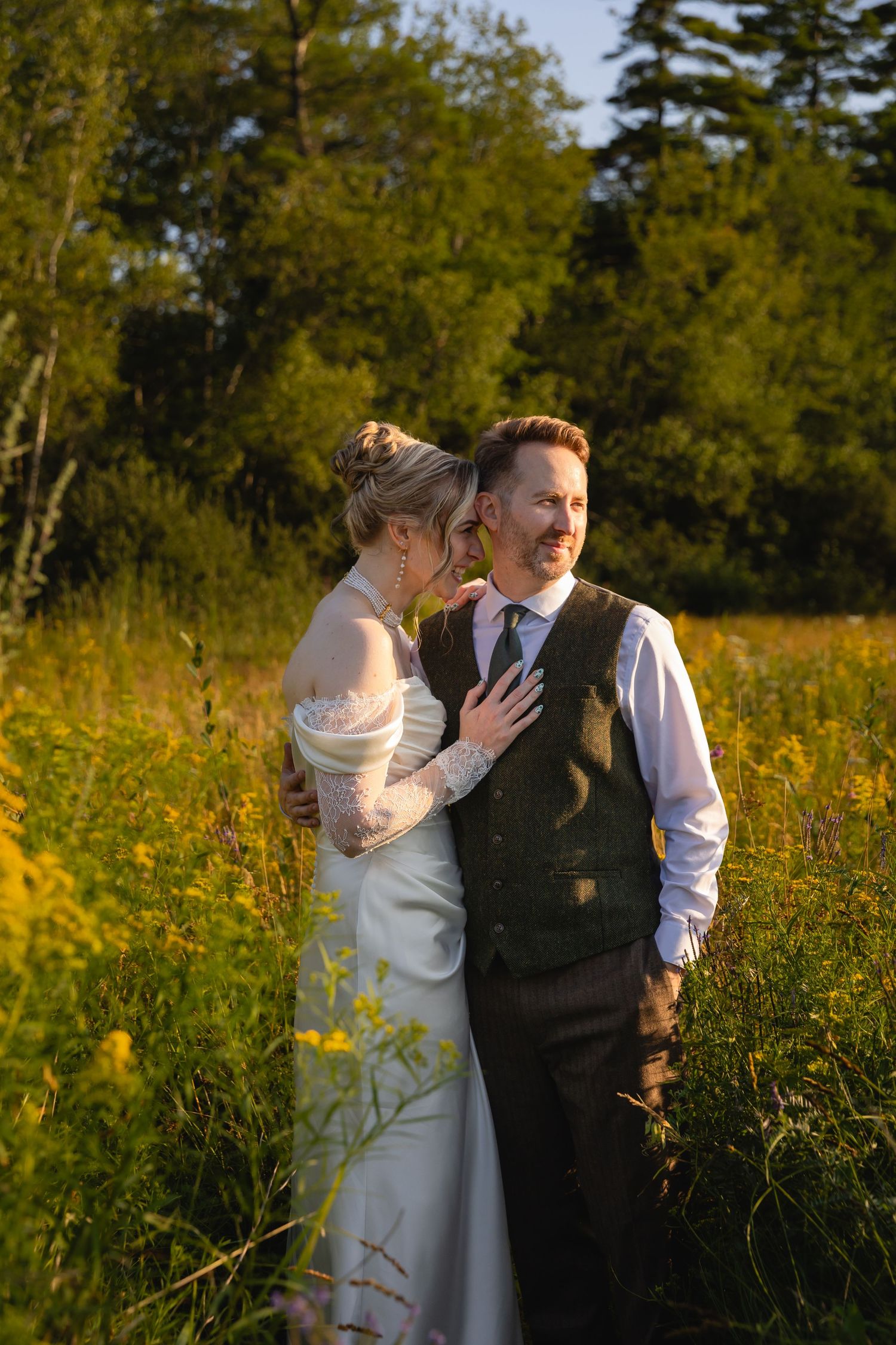 A couple shares intimate moments in a golden meadow during sunset surrounded by wildflowers and evergreen trees.