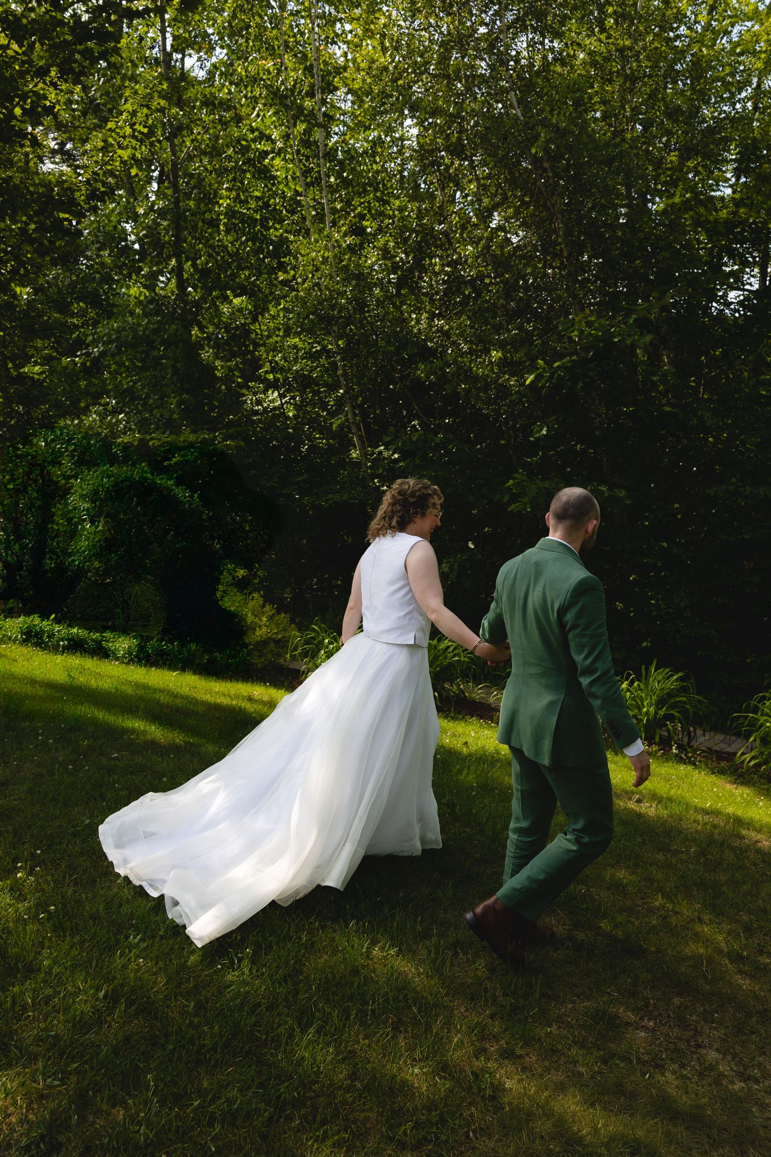 A couple in wedding attire walks through a sunlit garden path with flowing white dress and green suit.