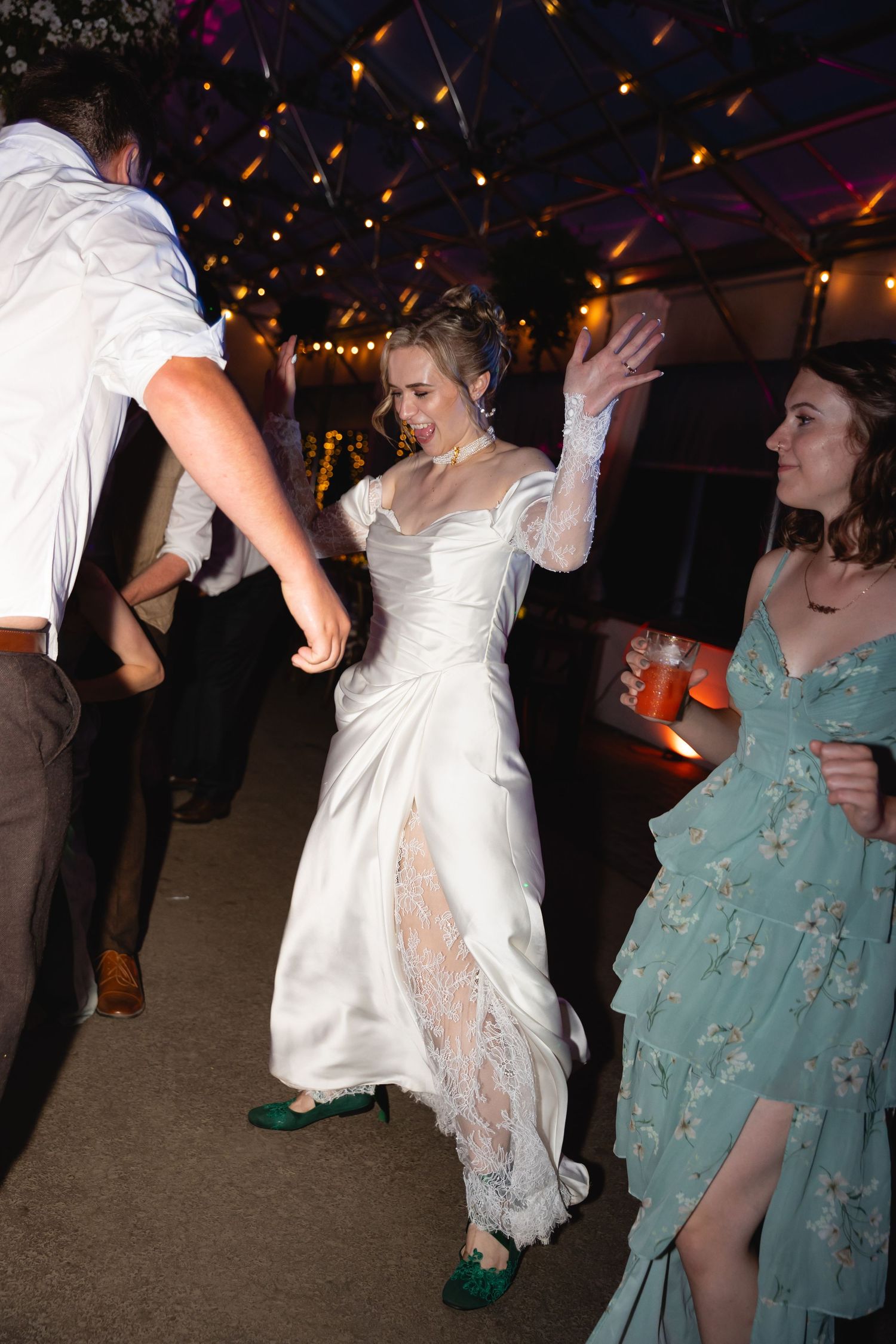 People dancing at a wedding reception with string lights overhead and someone wearing a white dress and teal dress.
