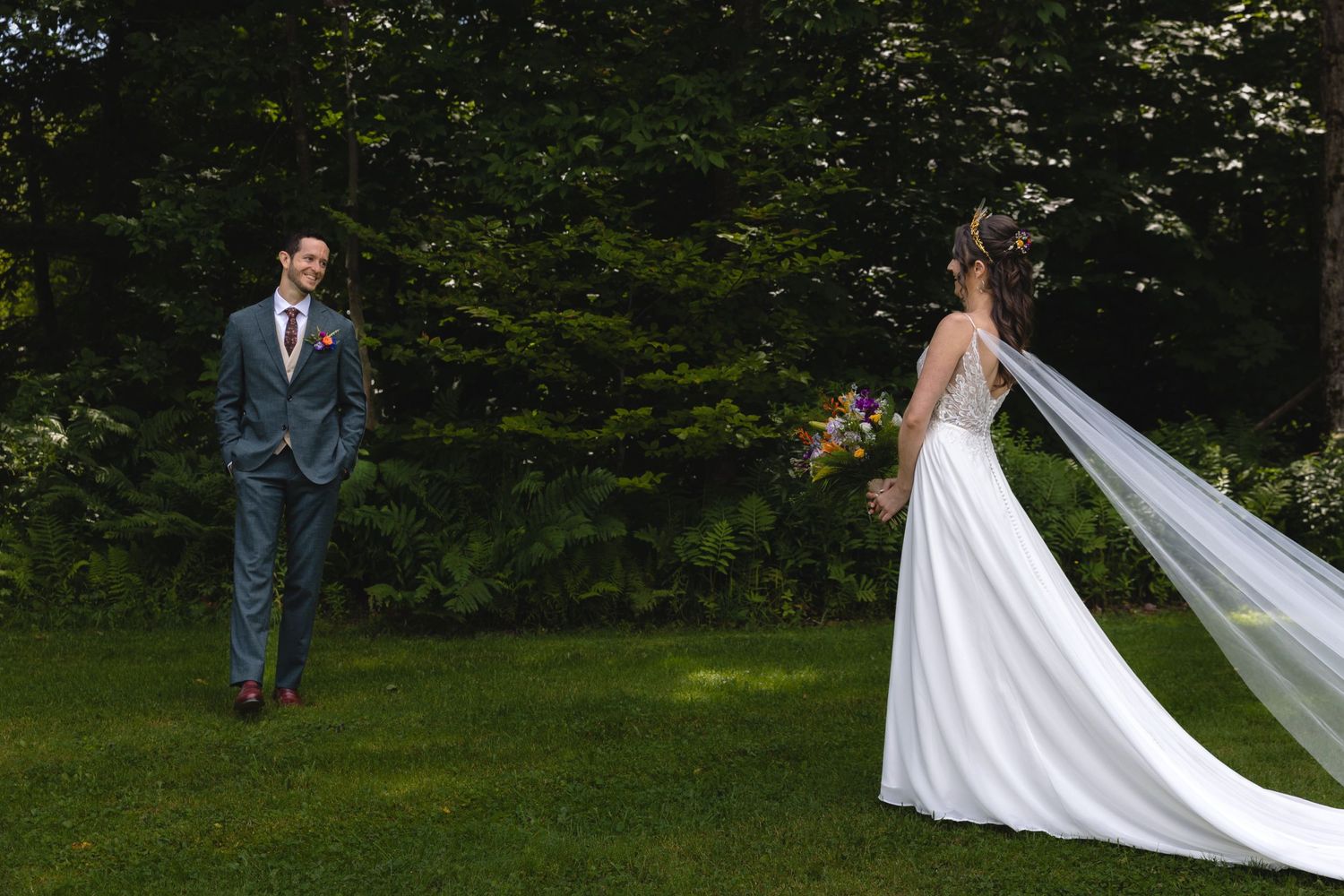 Wedding couple in elegant attire standing apart on green lawn with trees.