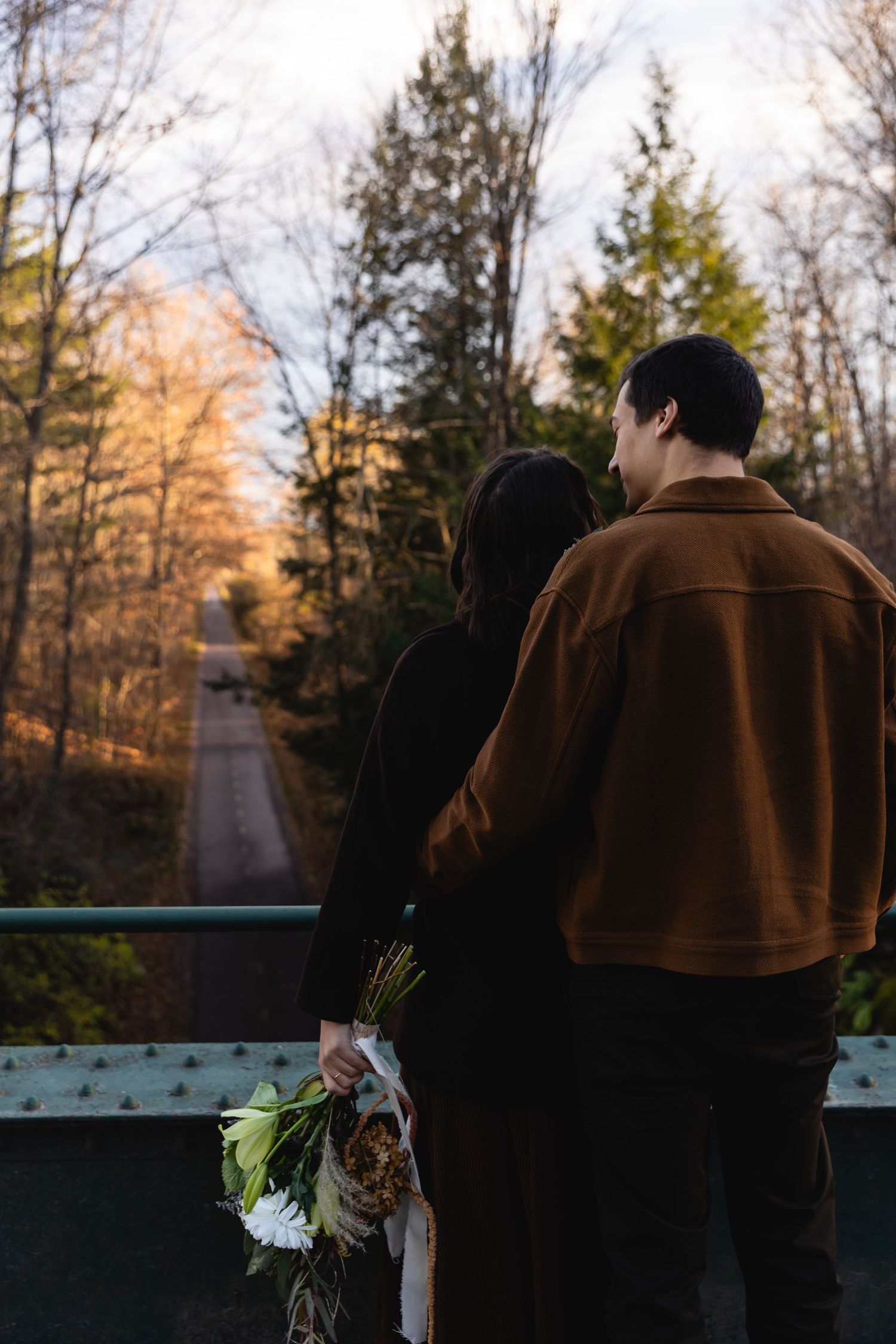 A couple stands on a bridge overlooking a tree-lined path during sunset while holding a bouquet of flowers.