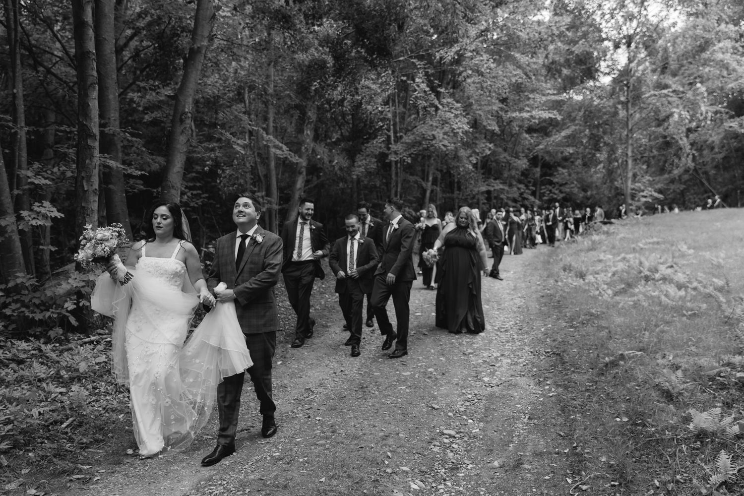 A wedding party walks in a procession down a wooded forest path in black and white.