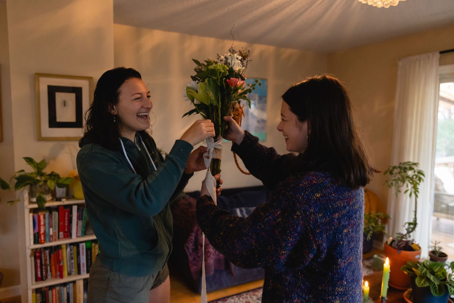 Two people playfully tugging at a bouquet of flowers in a cozy living room with bookshelves and natural light.