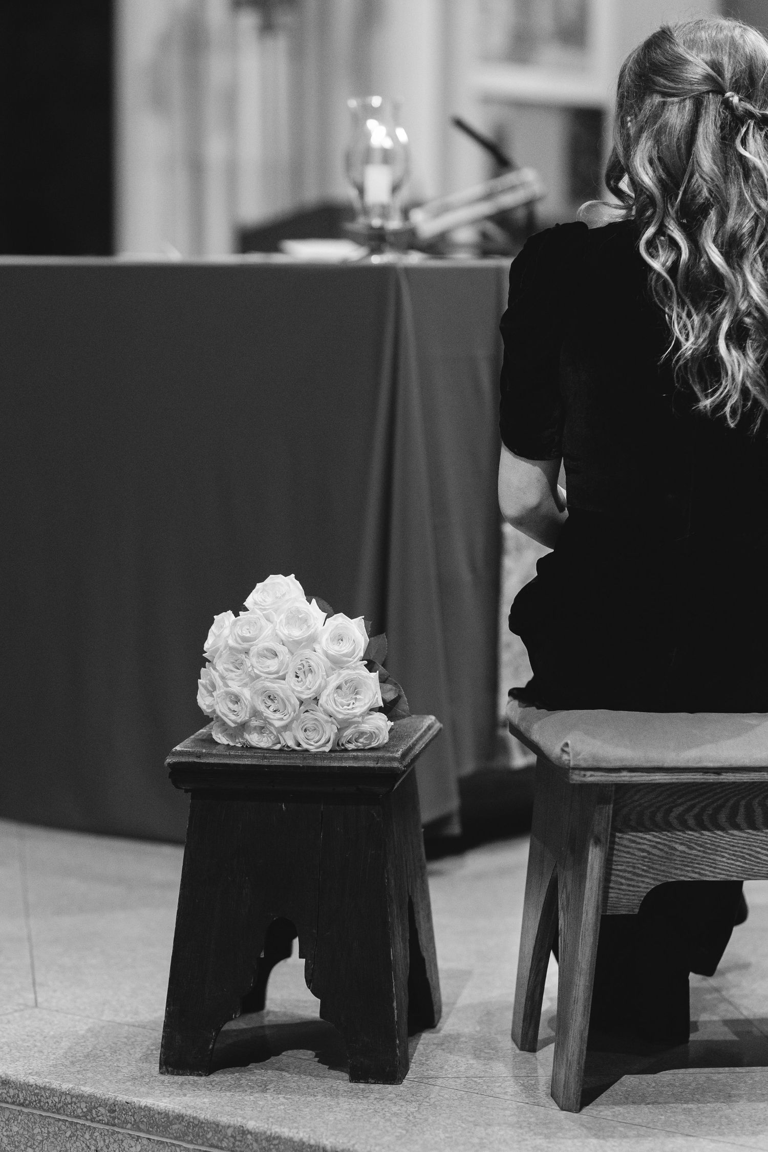 Black and white photo of a person sitting next to white roses on a small wooden stool during a formal event.