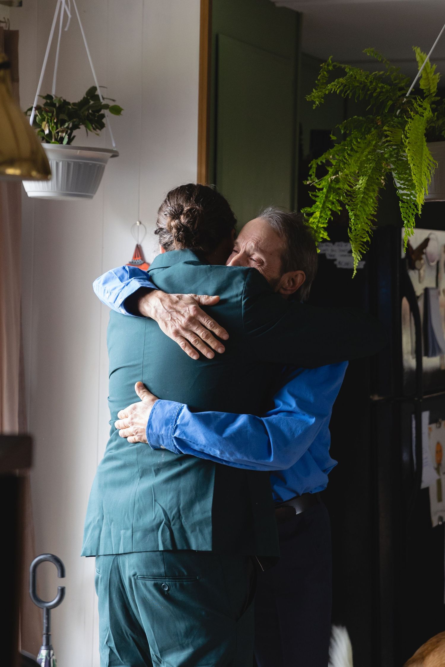 Emotional embrace between two people wearing teal and blue in an indoor setting with plants.