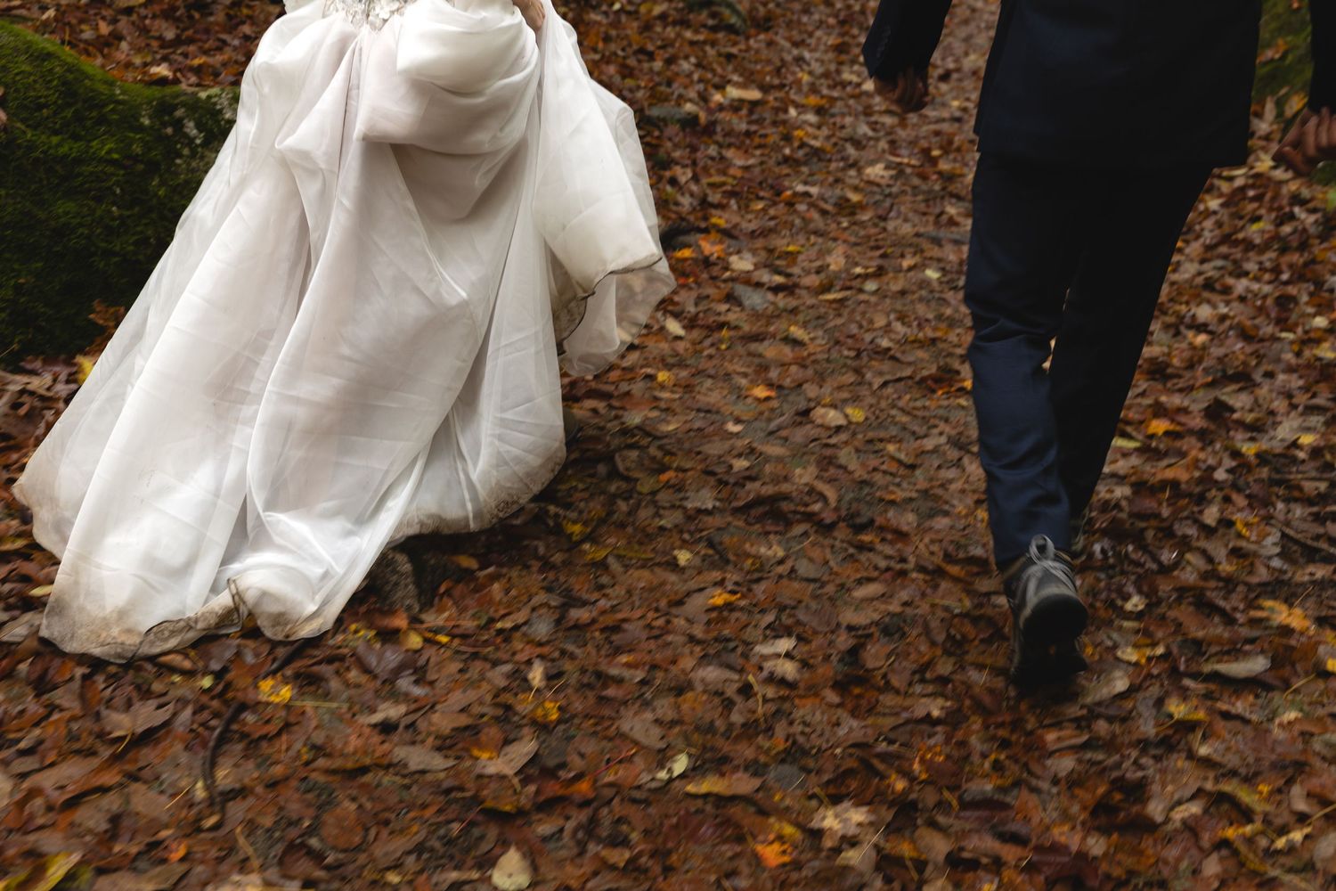 Wedding dress trailing through autumn leaves on a forest path as couple walks.