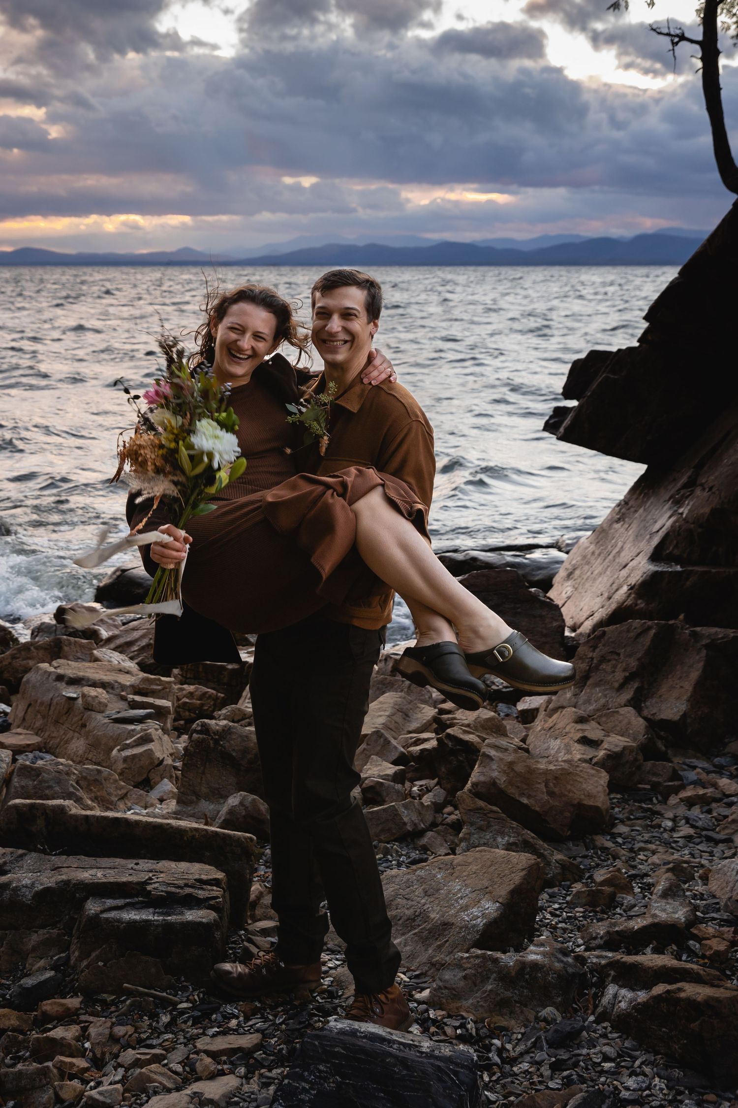 A couple enjoying a tender moment on rocky coastline with dramatic sunset and mountains in the background.