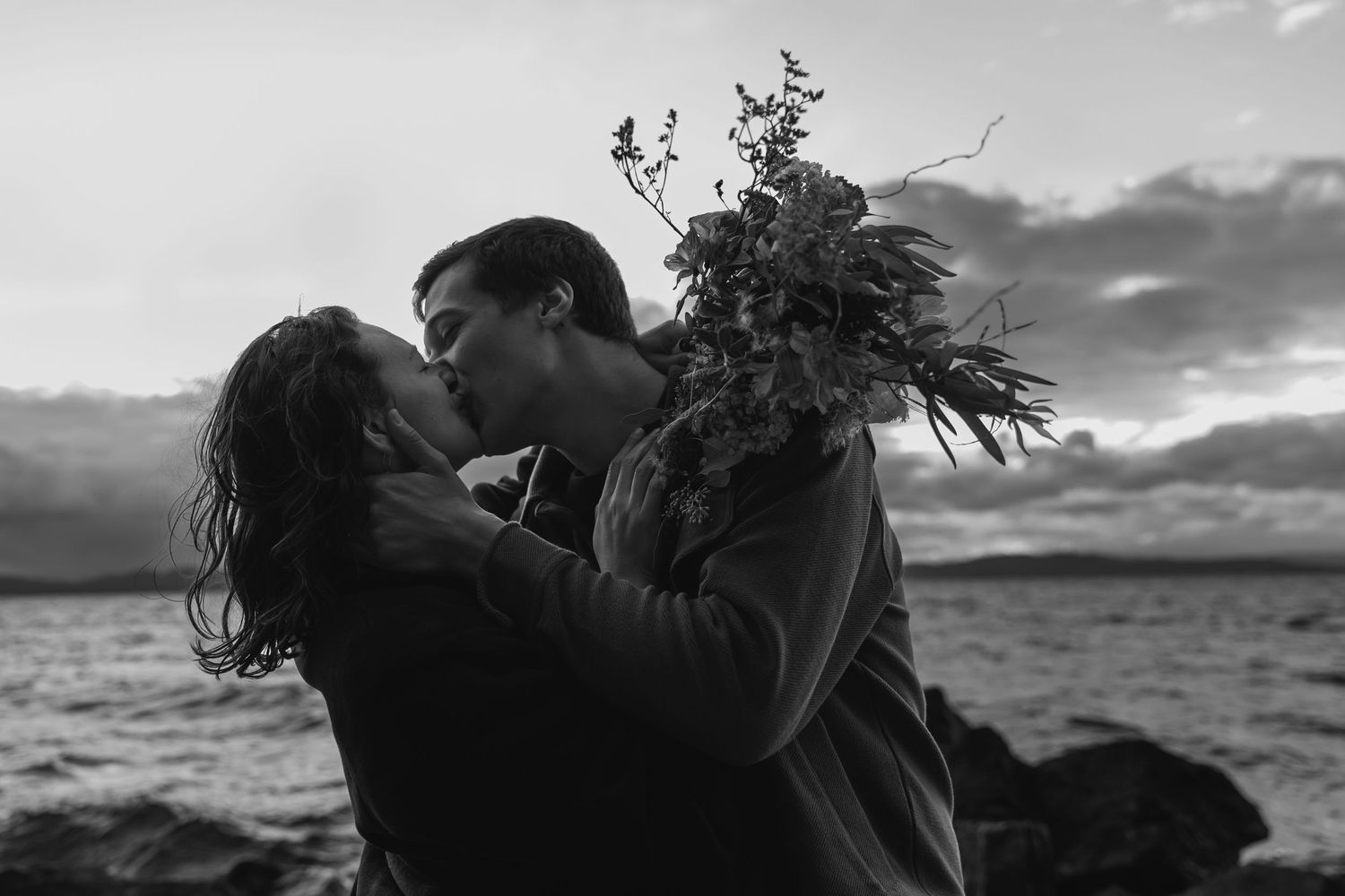A romantic silhouette couple shares an intimate moment by a lake at sunset while holding wildflowers.