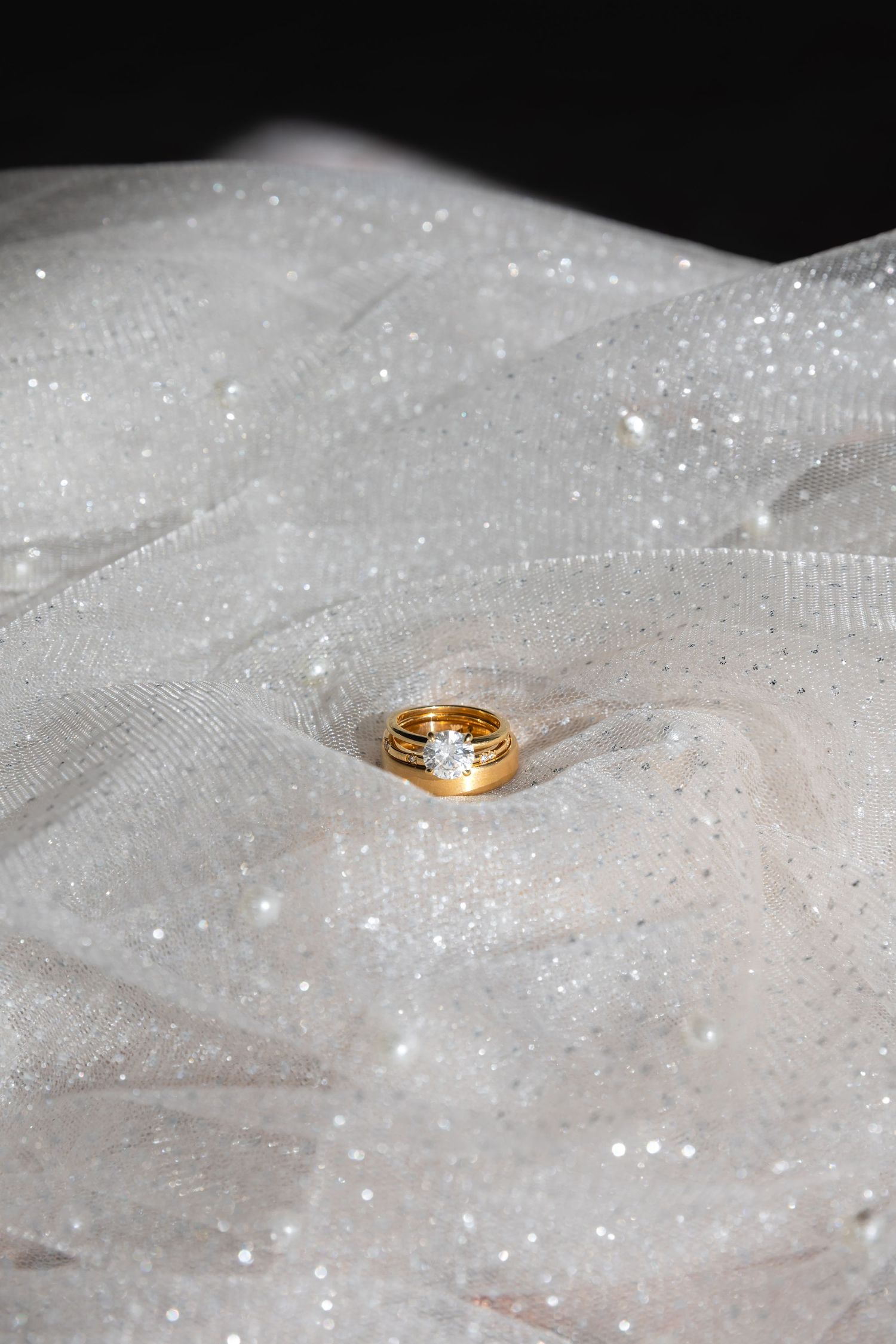 Gold engagement ring with diamond solitaire rests on sparkly white tulle fabric in dramatic lighting.
