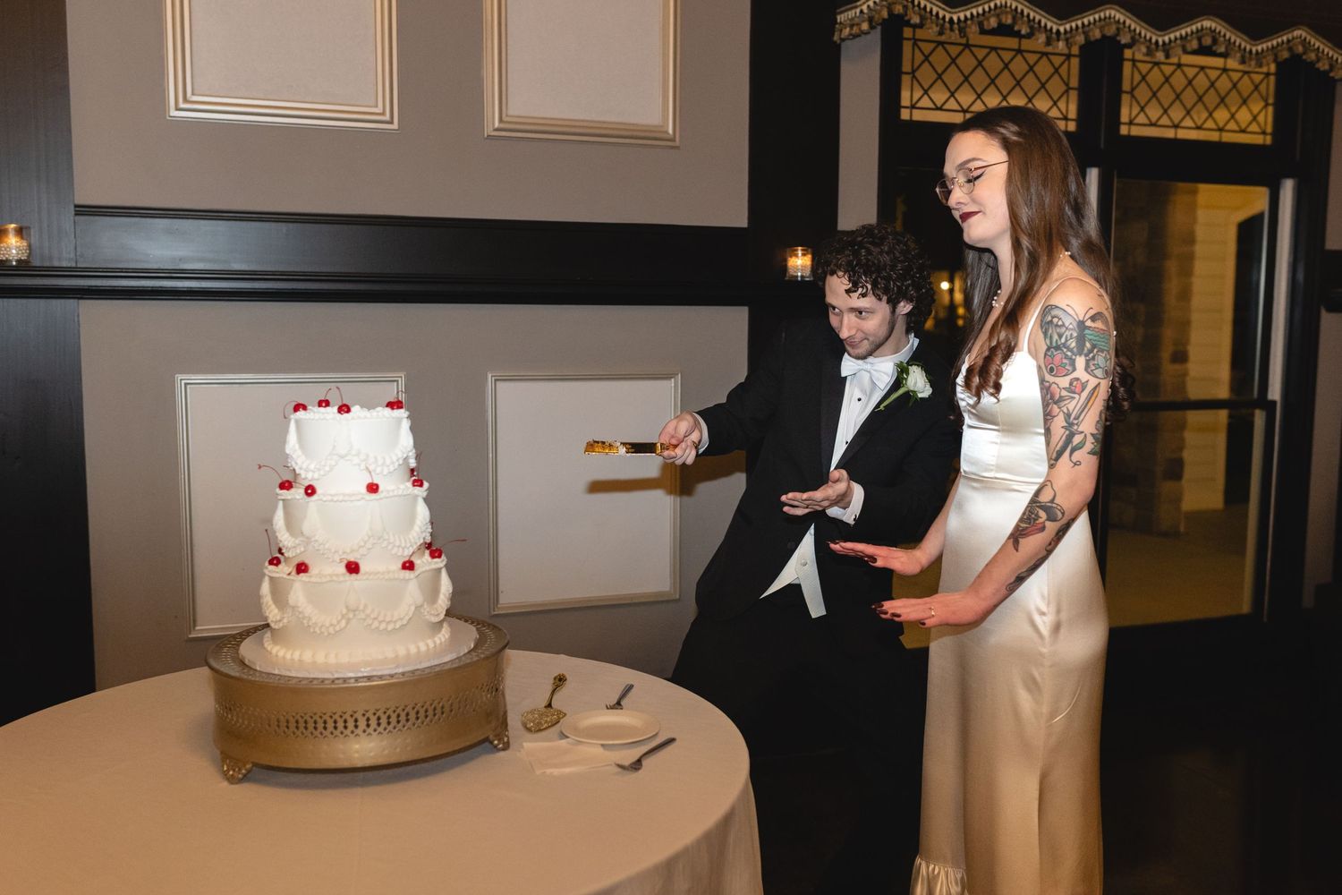 A couple at their wedding reception stands next to their white multi-tiered cake decorated with red accents.