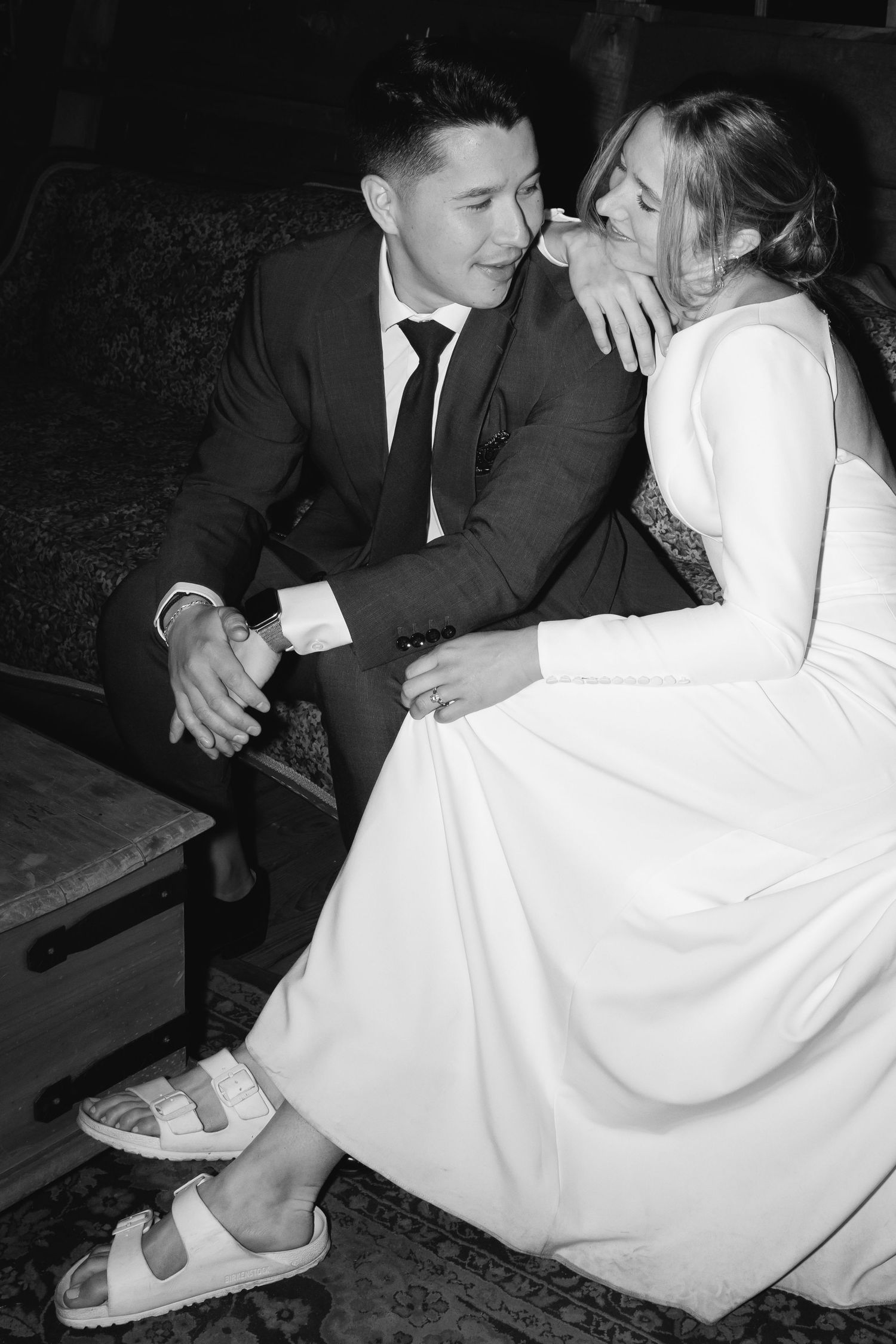 A black and white photo of a couple in wedding attire sharing an intimate moment on a vintage couch.