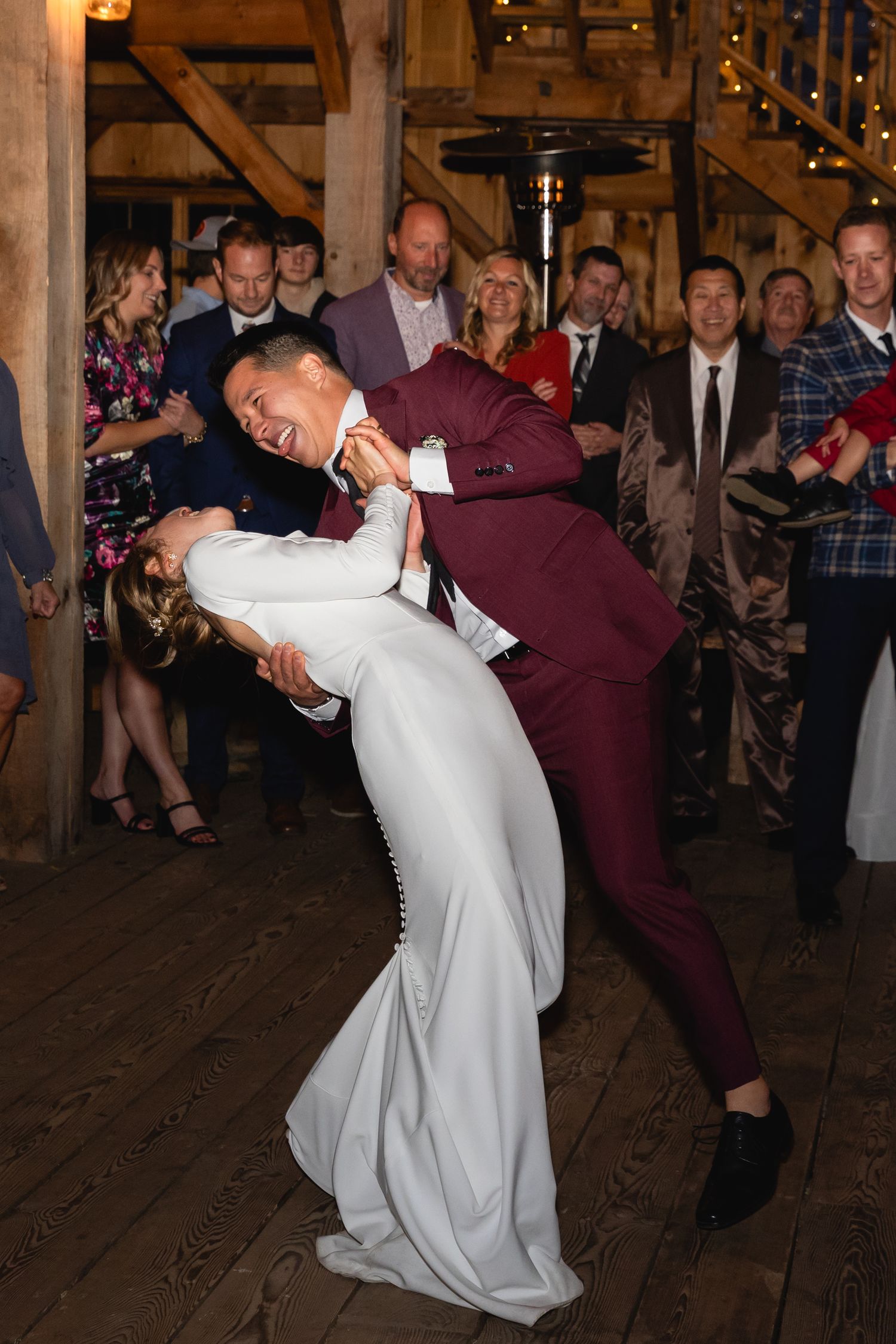 A joyful couple dips during their first dance at a rustic barn wedding reception with wooden beams and onlooking guests.