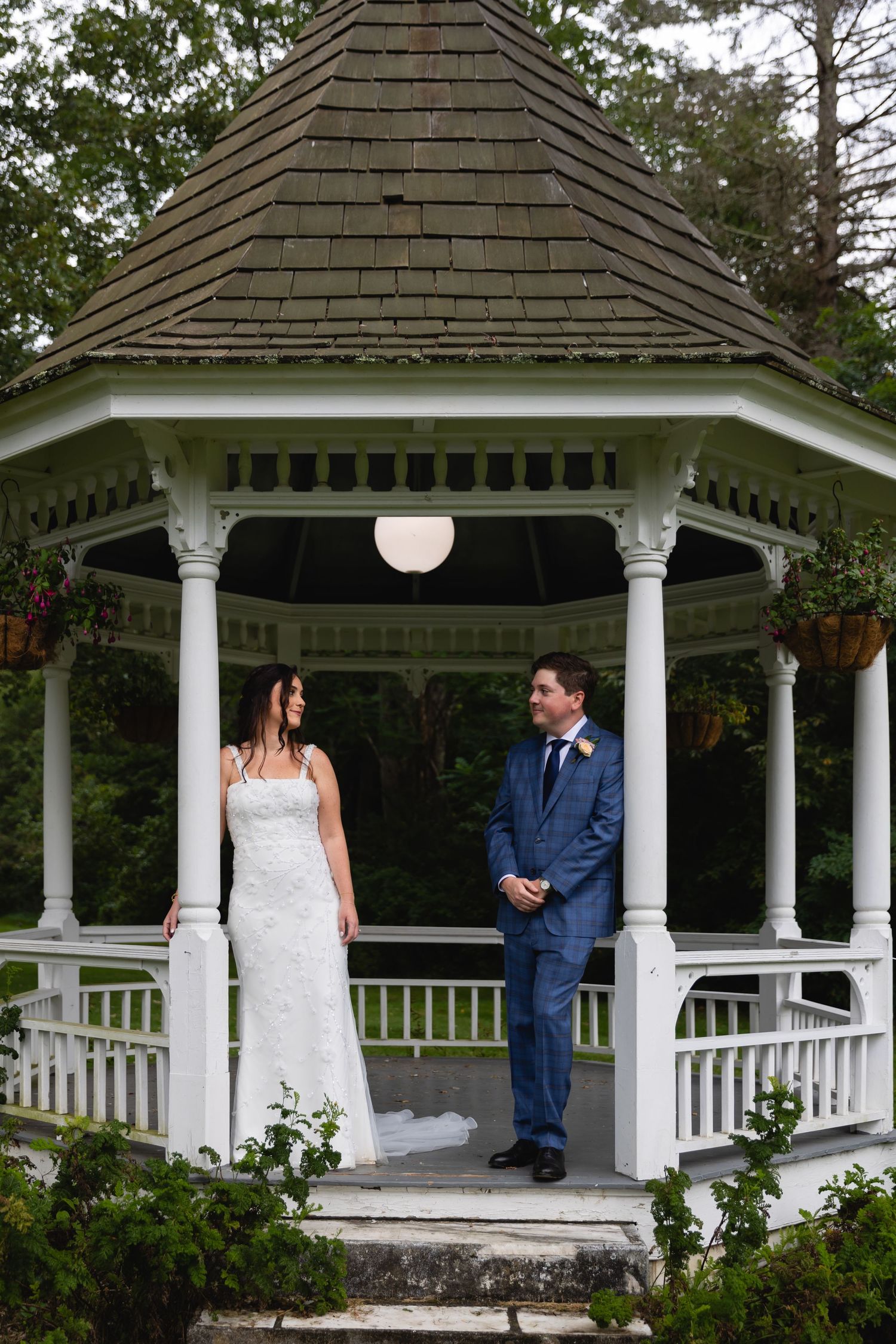 A white wooden gazebo with a shingled roof and decorative trim stands amid greenery, showcasing a romantic wedding scene.