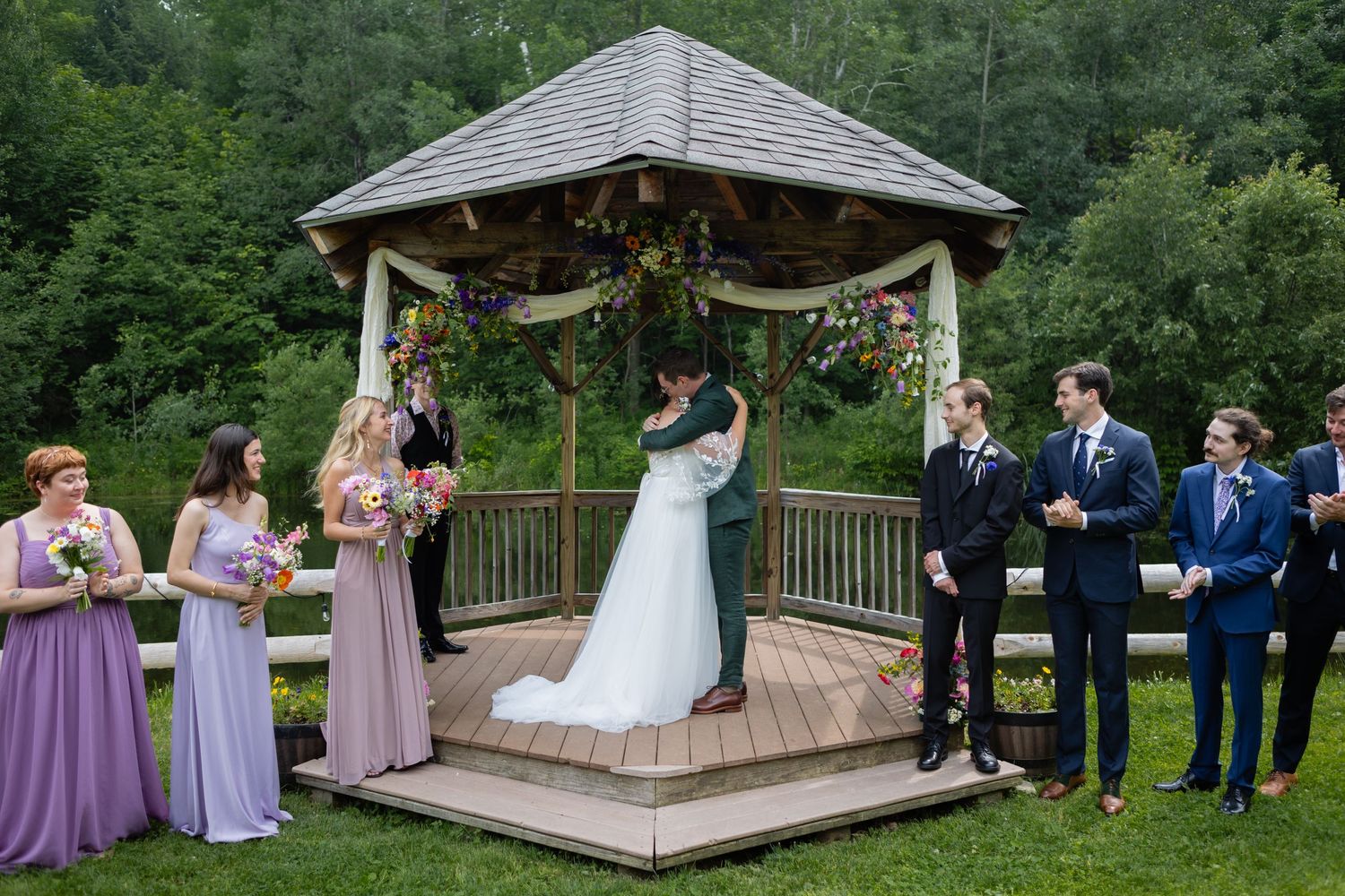 Full wedding party gathered during ceremony in gazebo decorated with purple flowers and fabric draping.