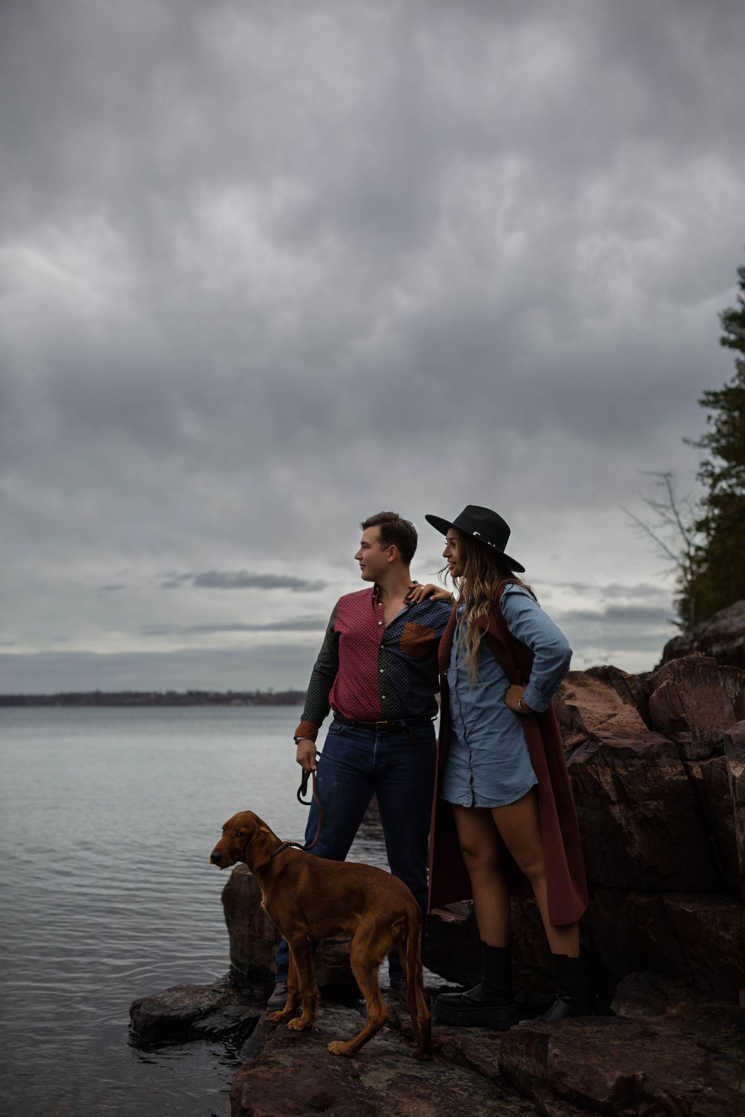 A couple and their dog stand on rocky cliffs overlooking a moody lakeside landscape.