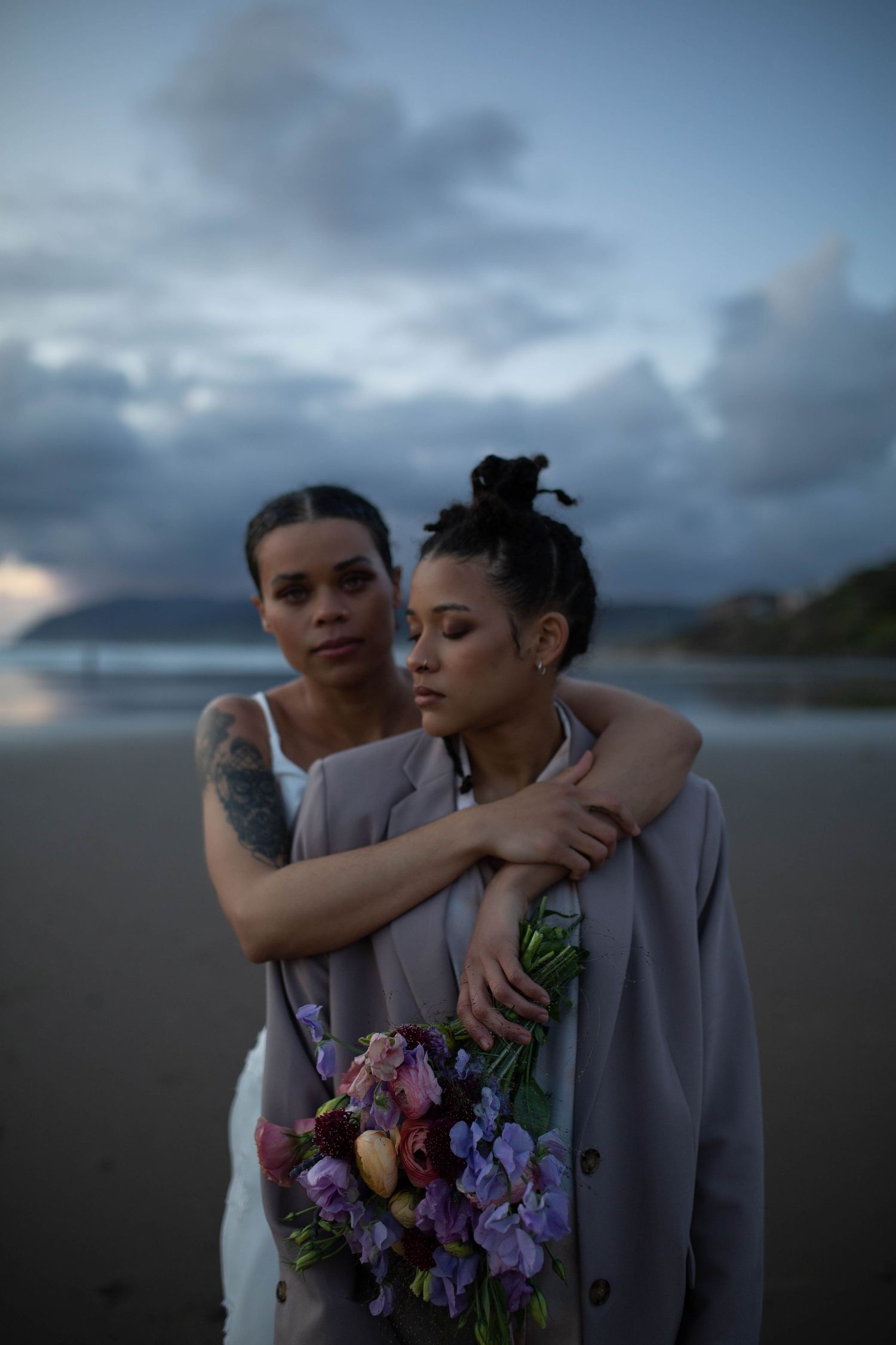 A couple shares an intimate embrace on a beach at dusk while holding colorful flowers.
