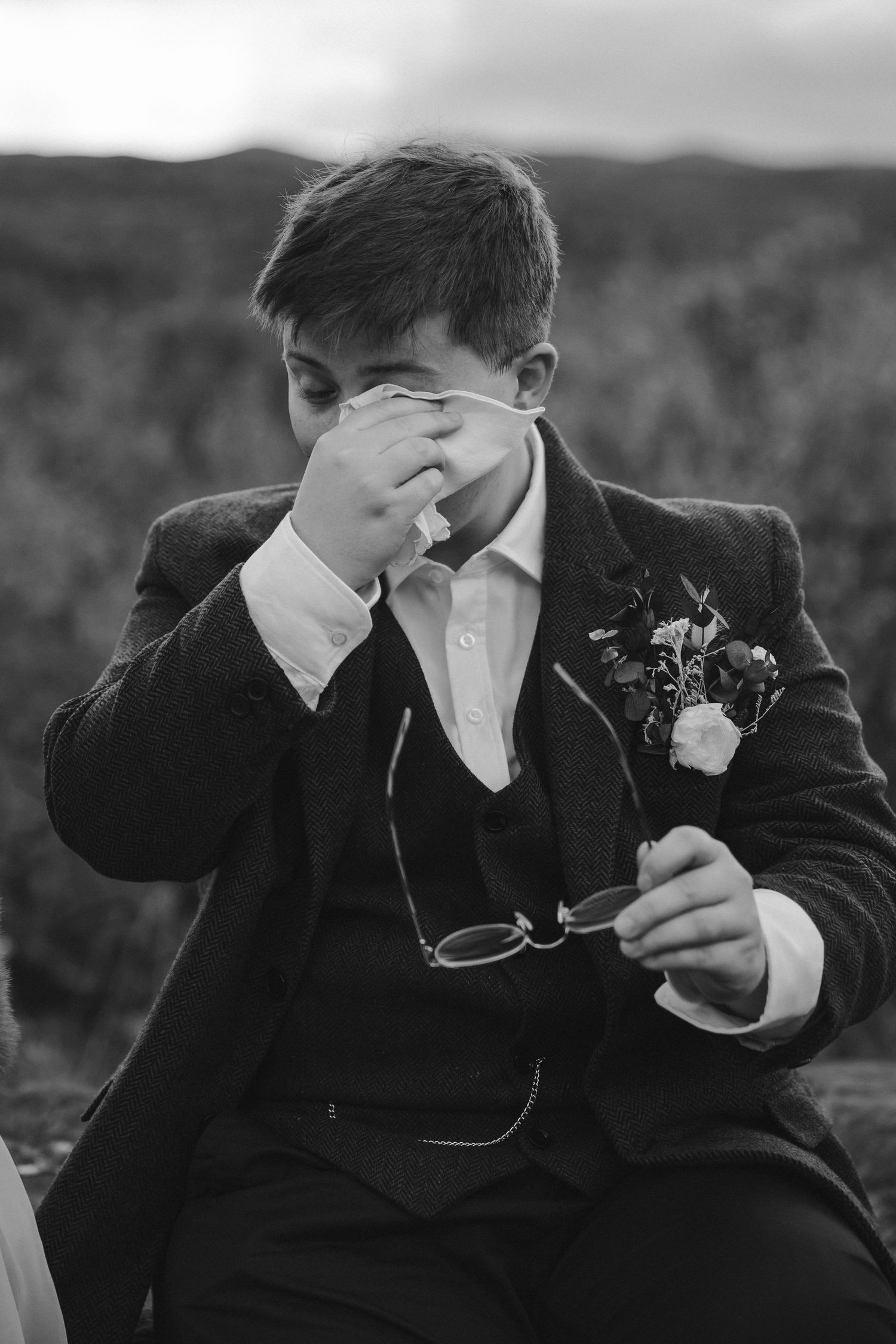 Black and white portrait of person in suit holding glasses with mountain backdrop.