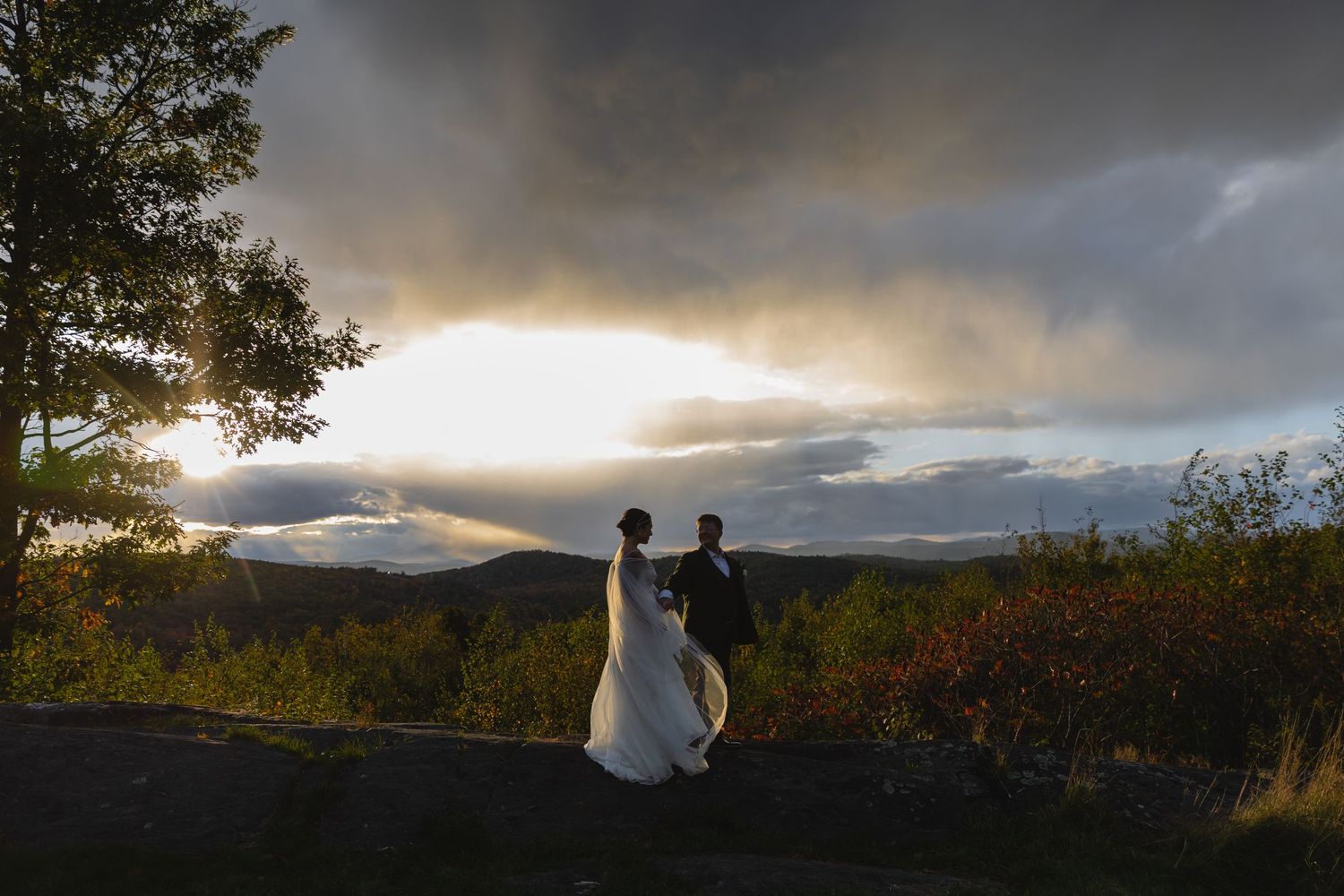 Silhouetted wedding couple stands on overlook with dramatic sunset sky and autumn foliage.