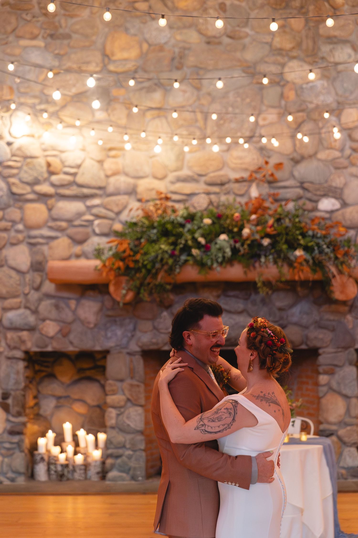 A couple shares an intimate dance moment in front of a stone fireplace with string lights.