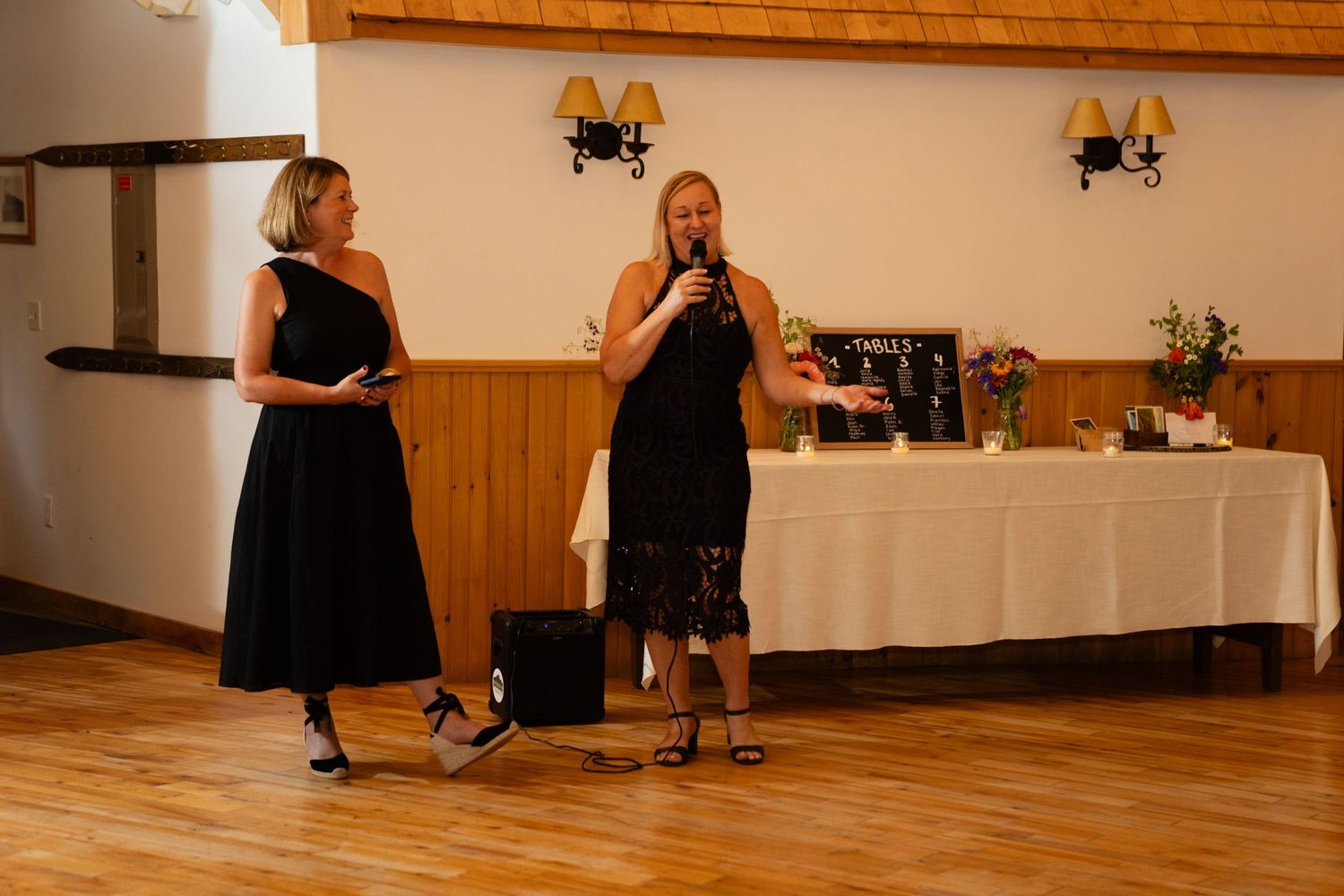 Two women in black dresses perform during wedding reception in rustic venue.