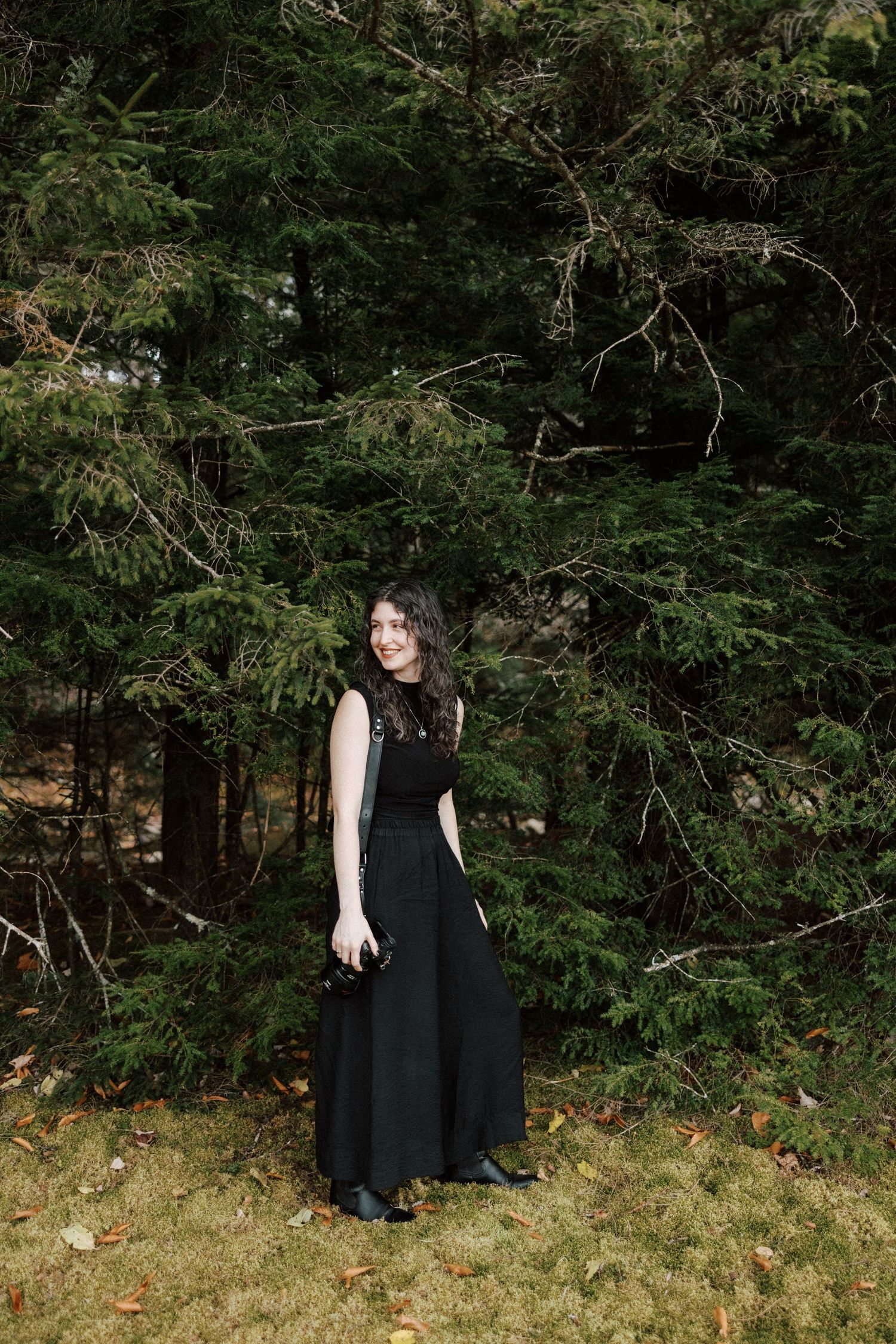 A smiling woman in a black sleeveless maxi dress stands before tall evergreen trees in a forest.