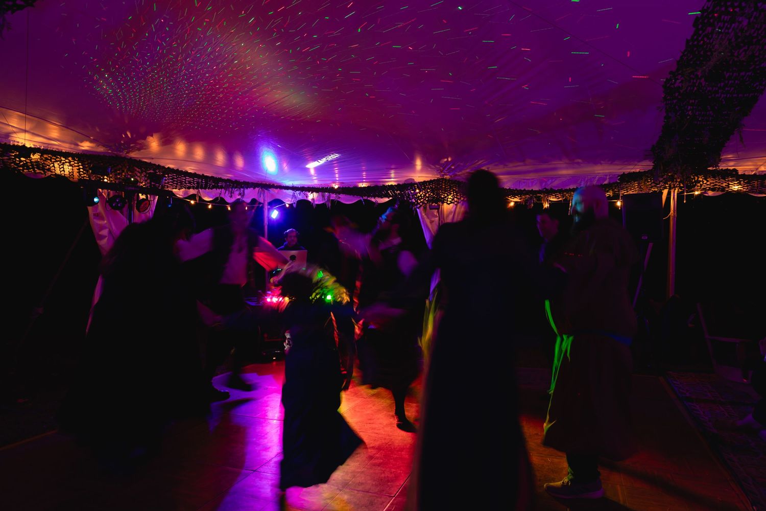 Crowd of wedding guests dancing under colorful laser lights in a large decorated tent.