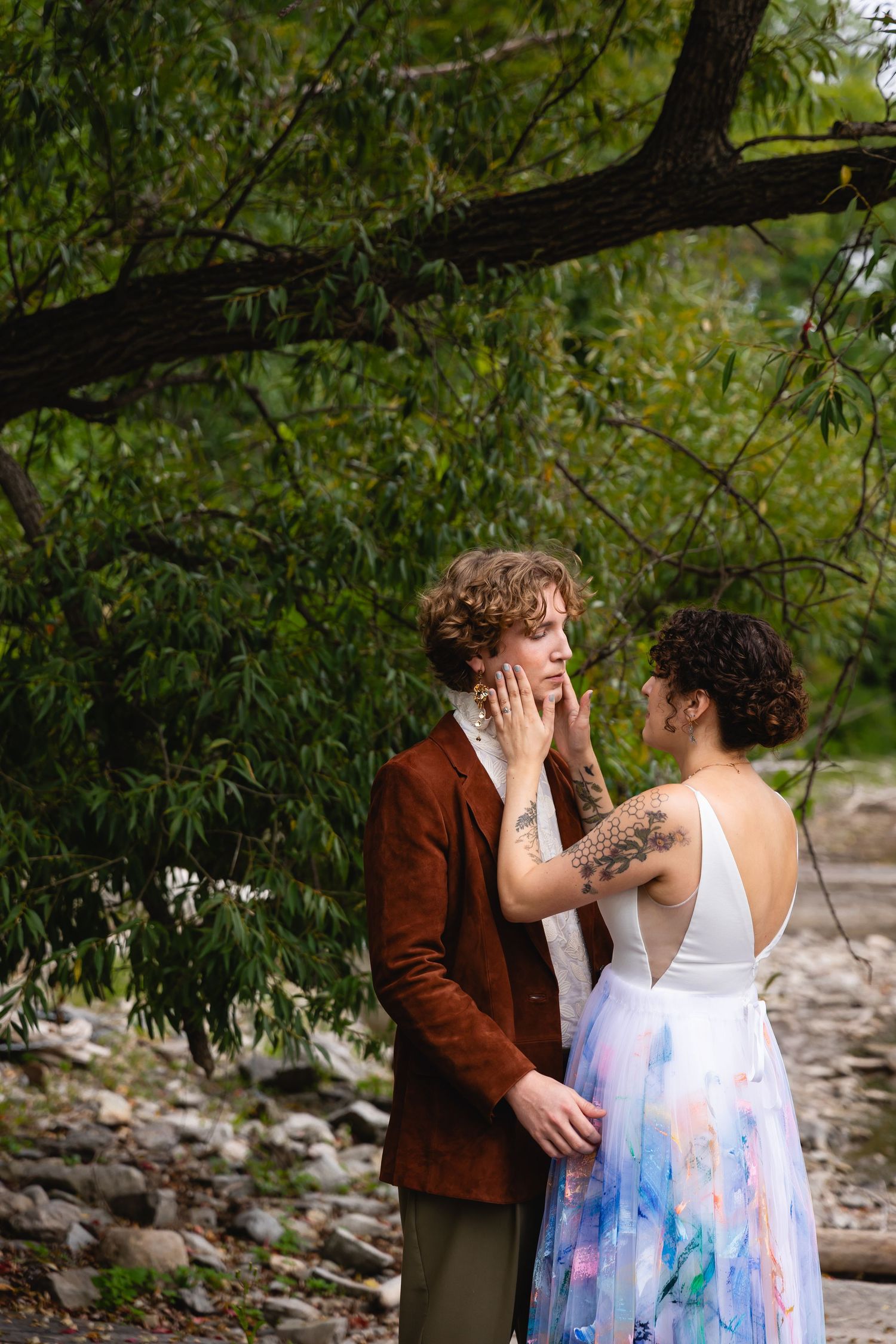 Couple sharing an intimate moment outdoors, woman in watercolor dress touching mans face near green trees.