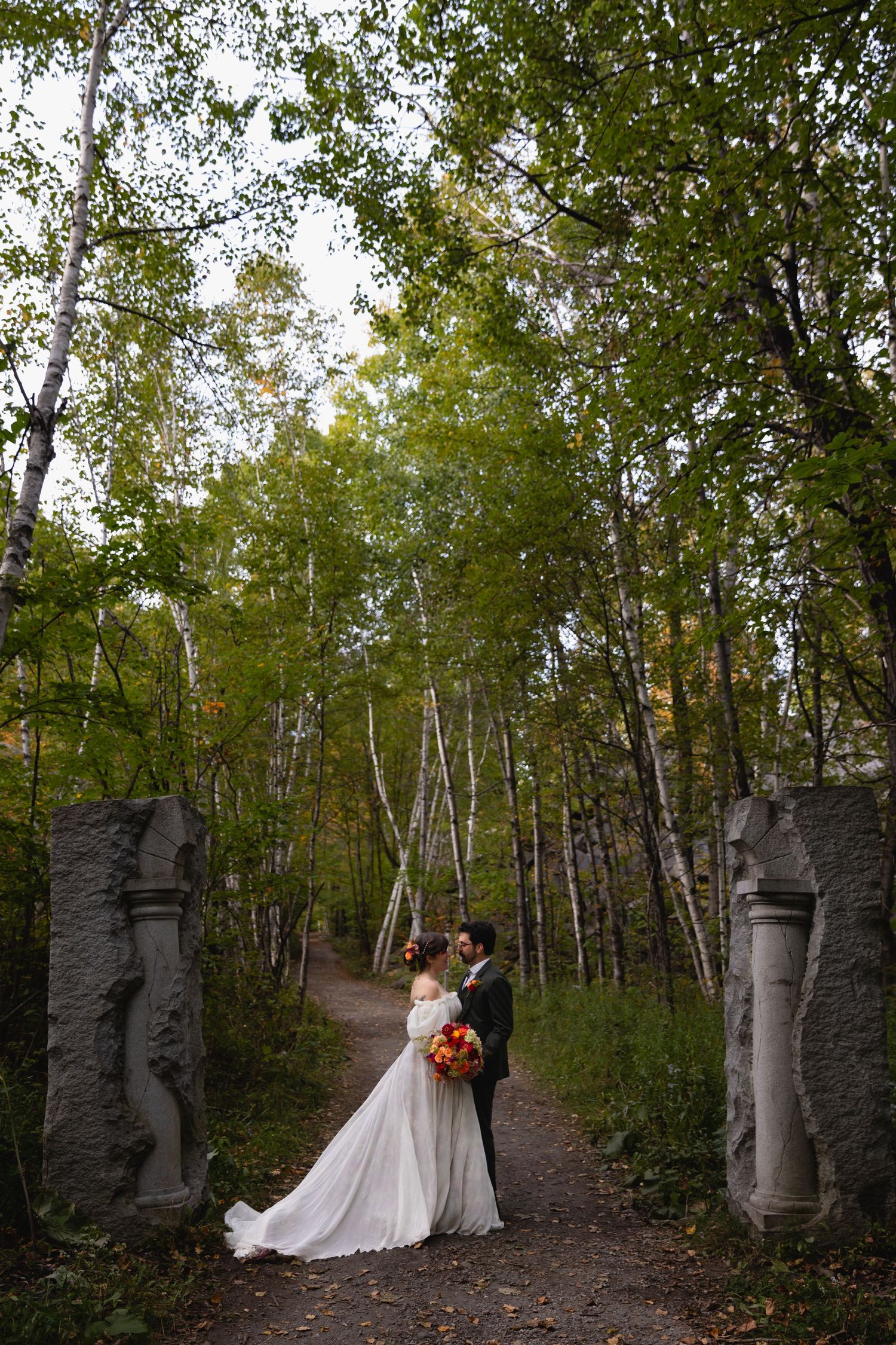 Bride and groom embrace between stone pillars on forest path with birch trees.