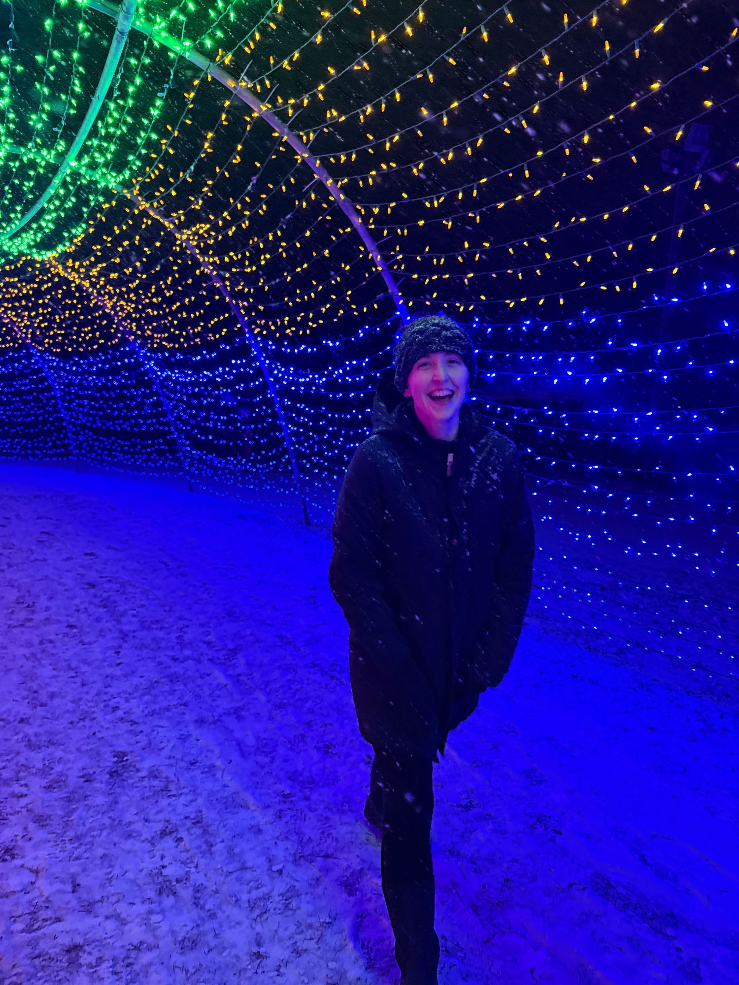 Person laughing joyfully while walking through colorful illuminated holiday light tunnel in snowy winter night.