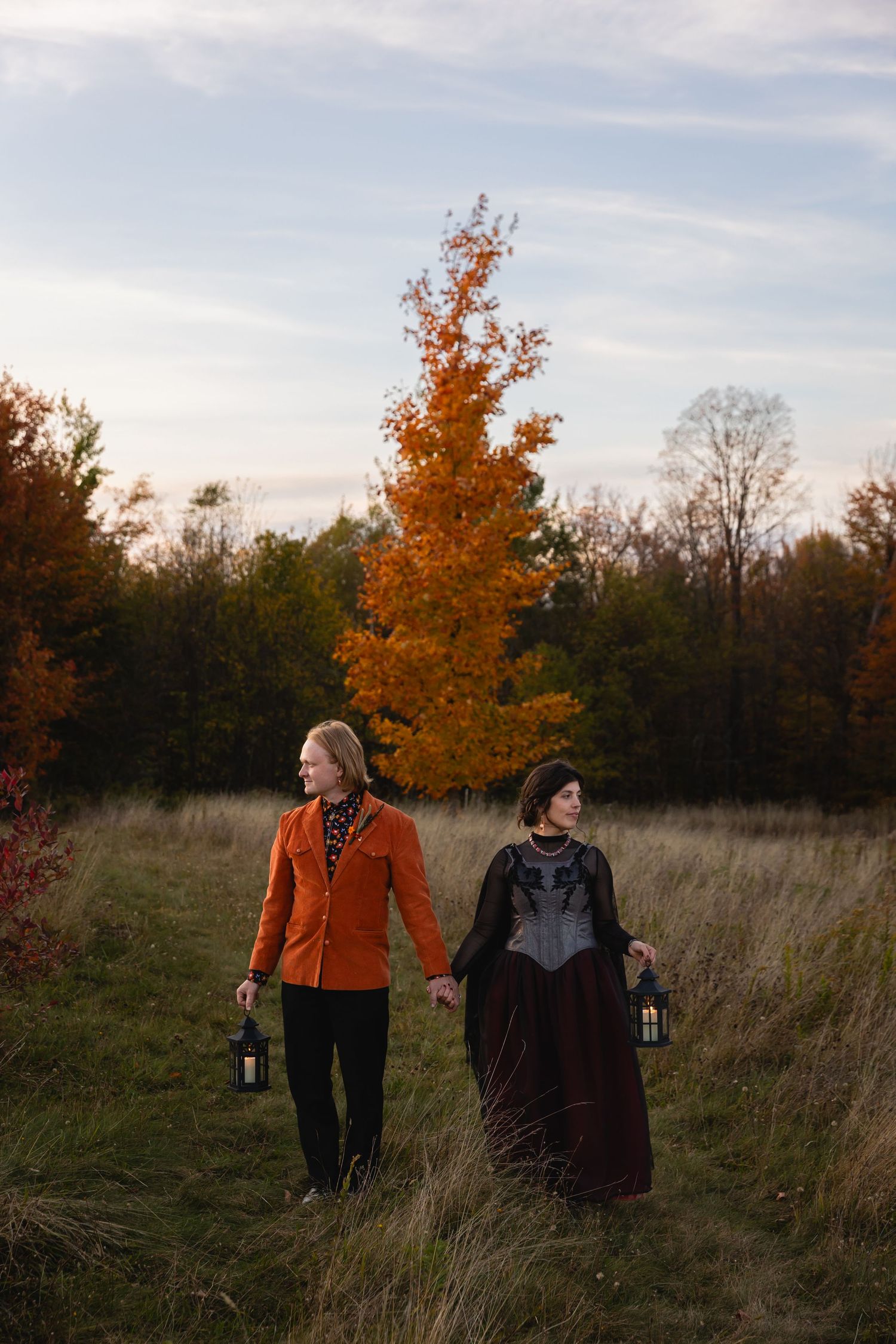 Couple in autumn field holding lanterns wearing orange blazer and dark corset dress.