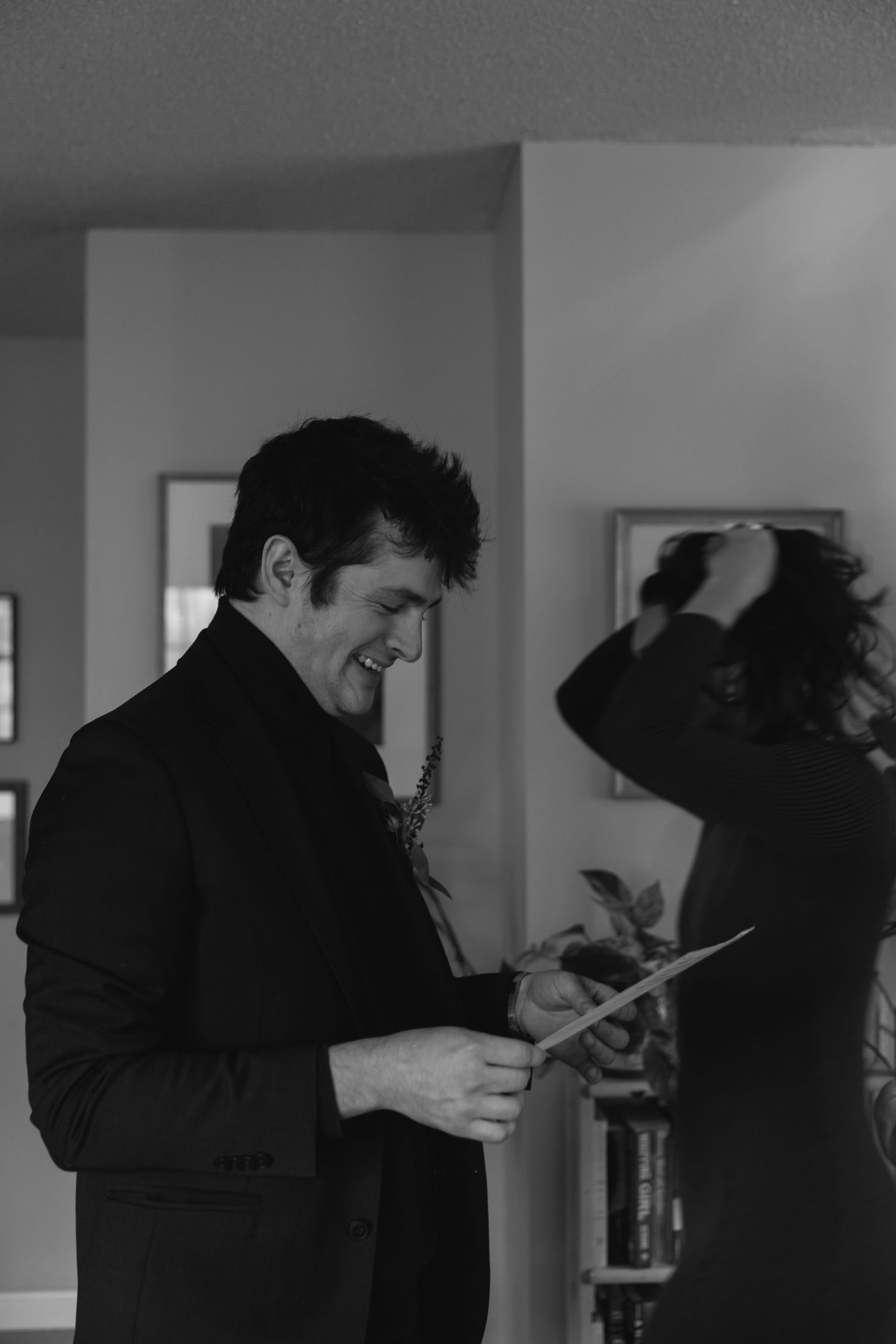 Black and white candid photo of someone in a dark turtleneck sweater smiling while reading something indoors.