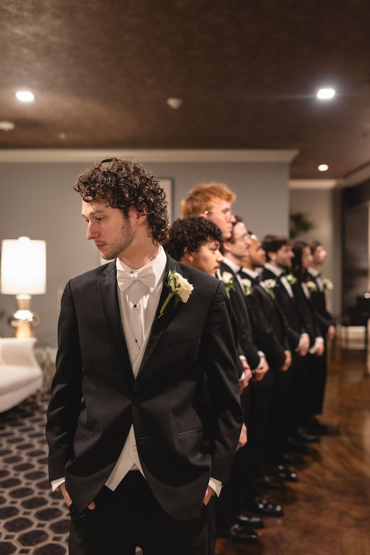 Groomsmen lineup in black tuxedos standing against a wall with warm lighting and elegant interior decor at wedding venue.