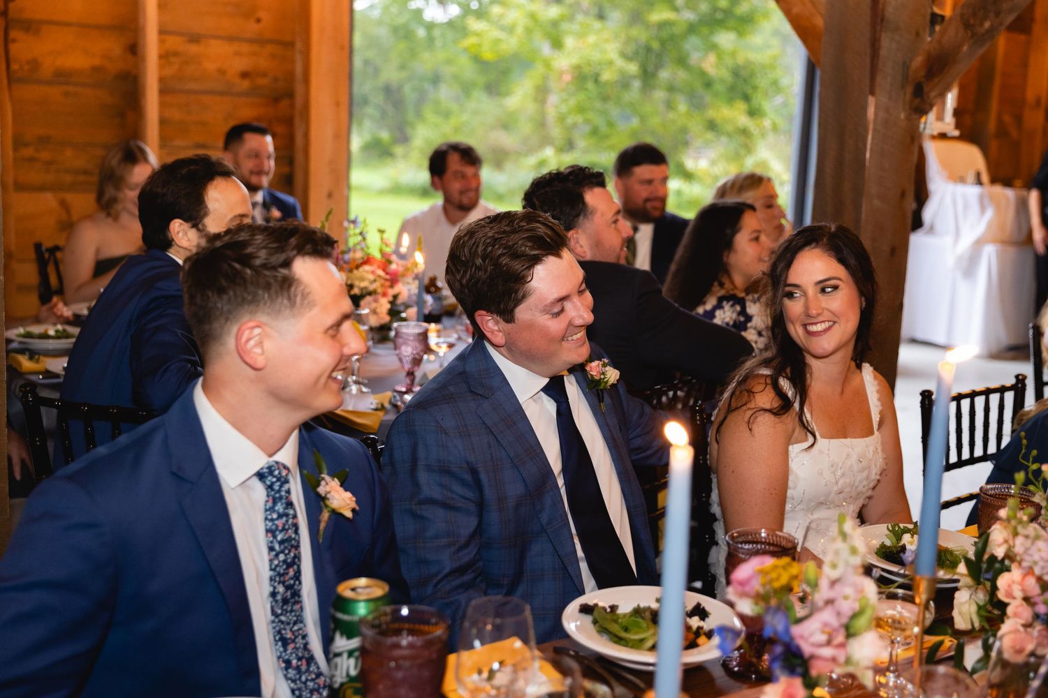 Wedding guests enjoy a meal and conversation at an elegant rustic barn reception with floral centerpieces.