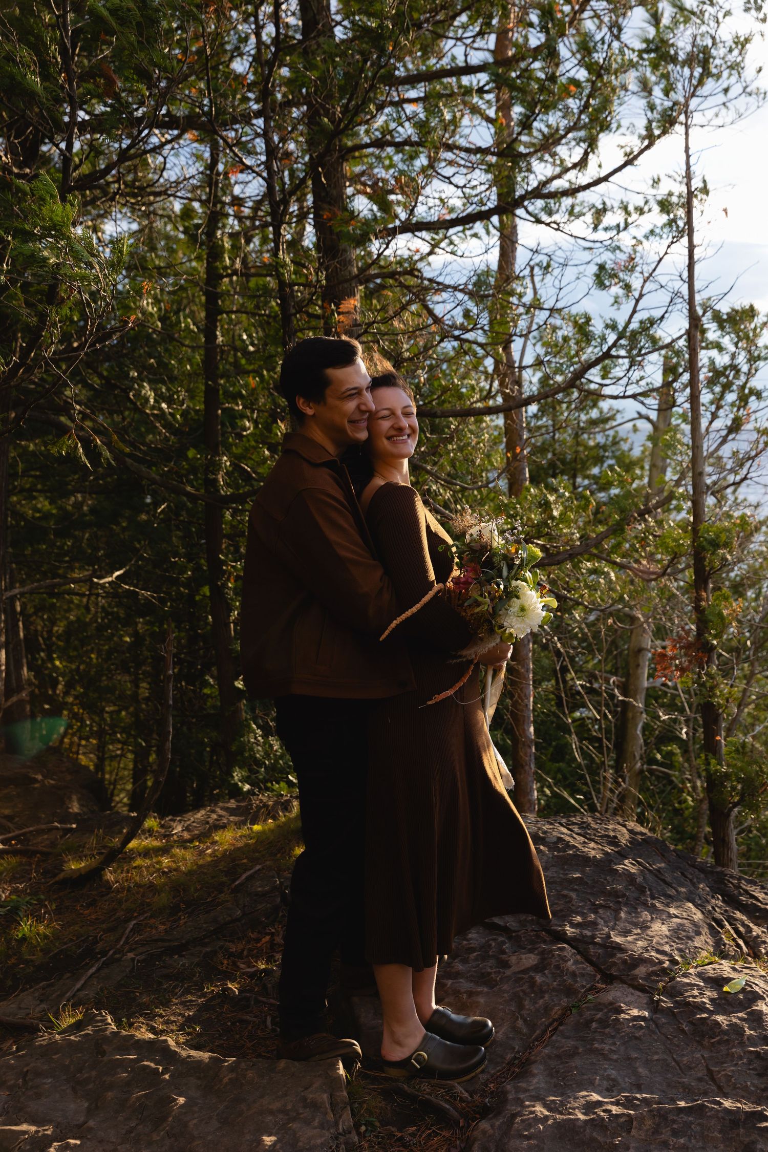 A couple embraces while standing on a rocky forest trail surrounded by tall pine trees on a sunny day.