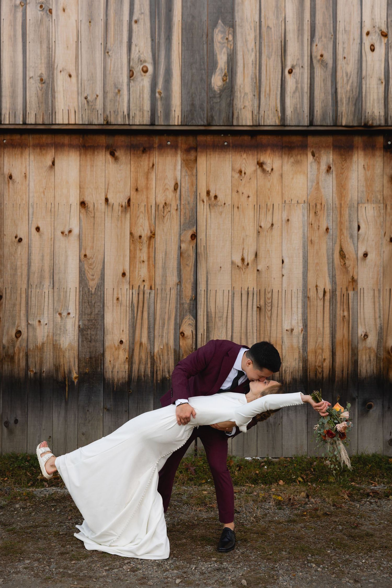 Couple sharing a romantic dip pose in front of rustic wooden barn wall with a wedding bouquet.