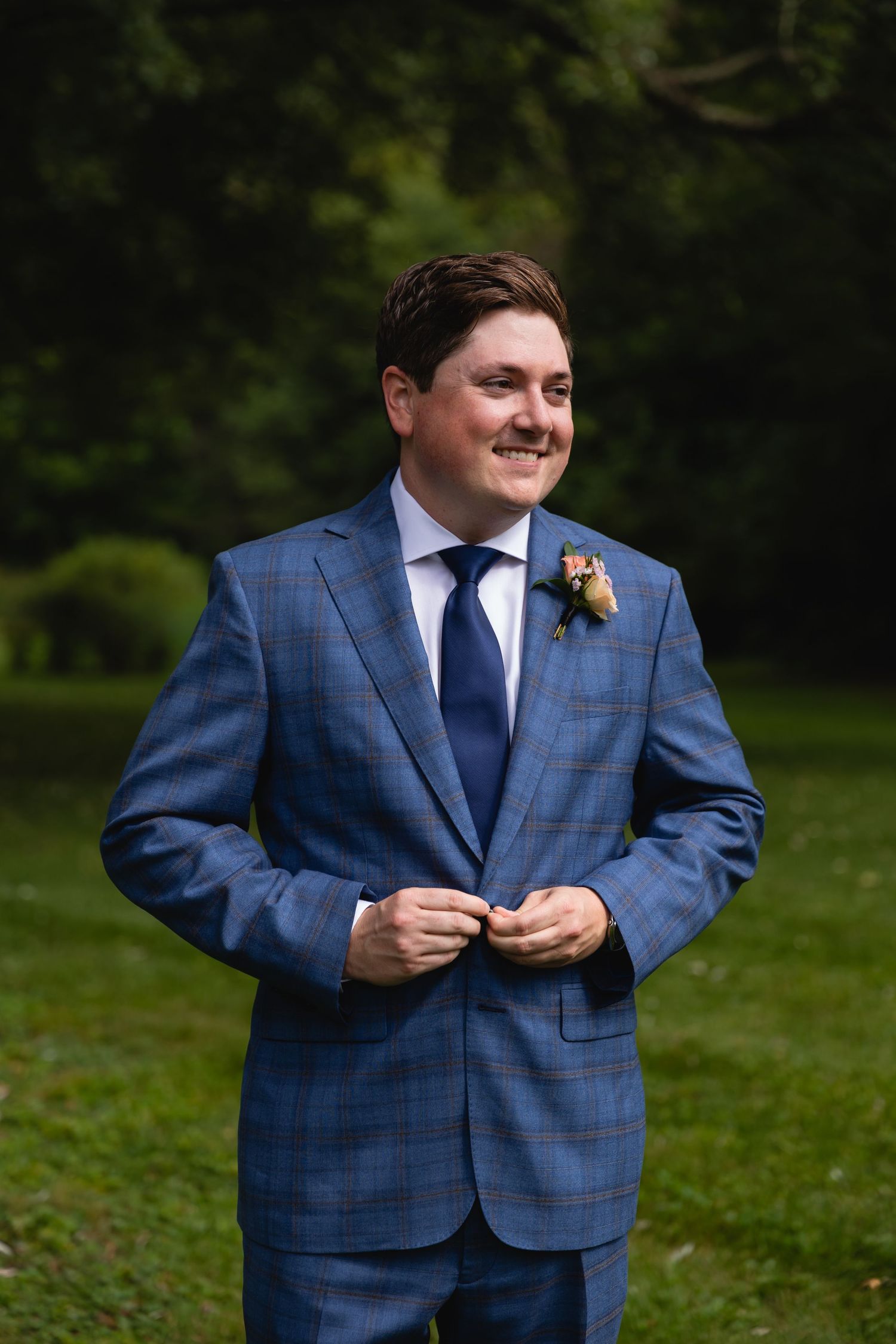 Groom in blue plaid suit jacket with boutonniere buttons his jacket while smiling outdoors.