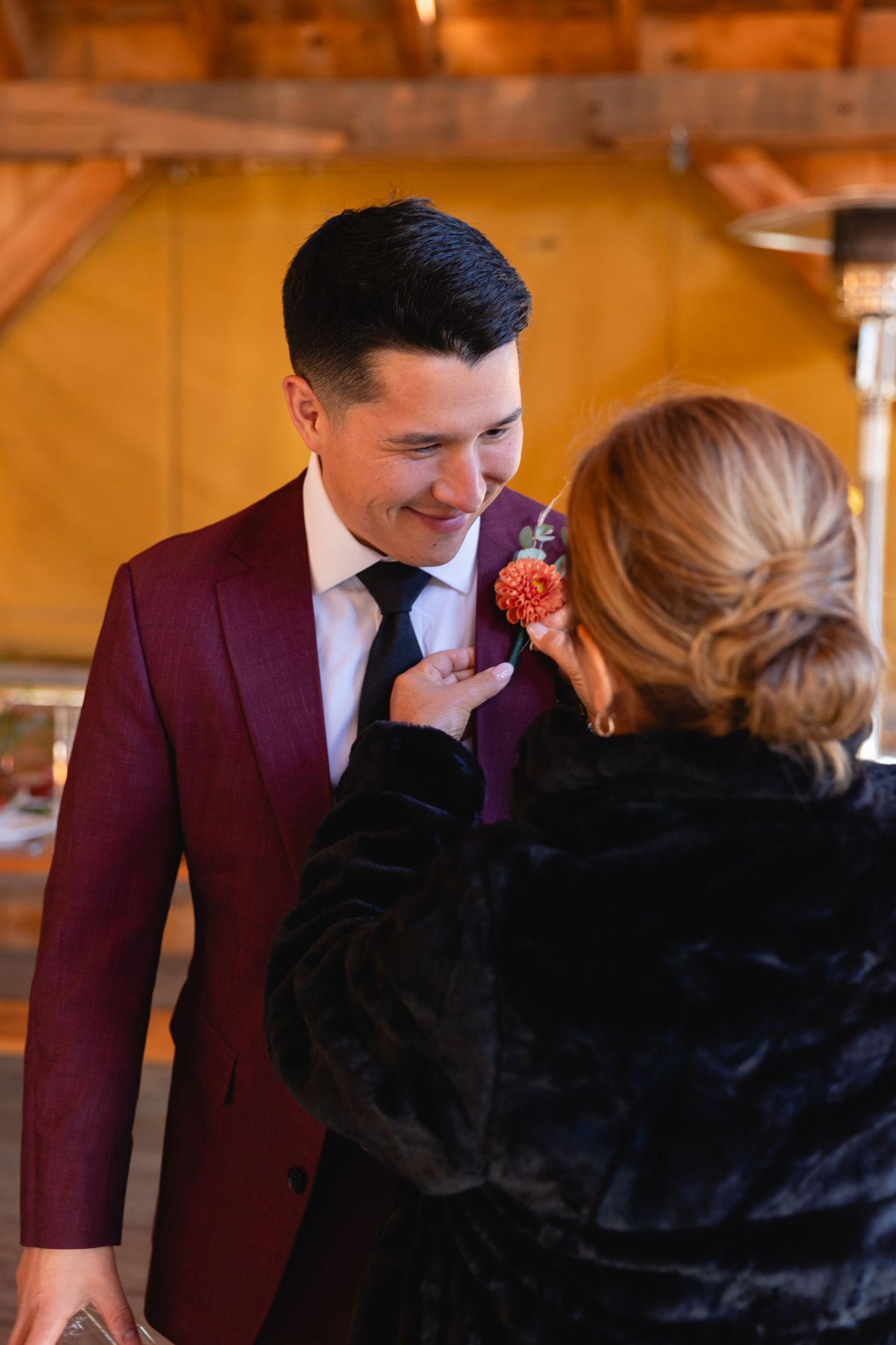 A groom in a burgundy suit receives a boutonniere pinning from someone wearing a black fur coat at an indoor venue.