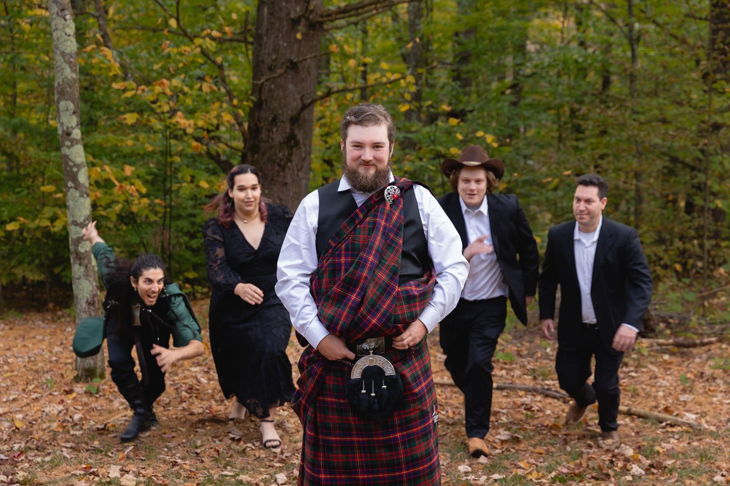 A wedding party poses playfully in a forest setting with fall foliage and a person wearing traditional Scottish kilt attire.