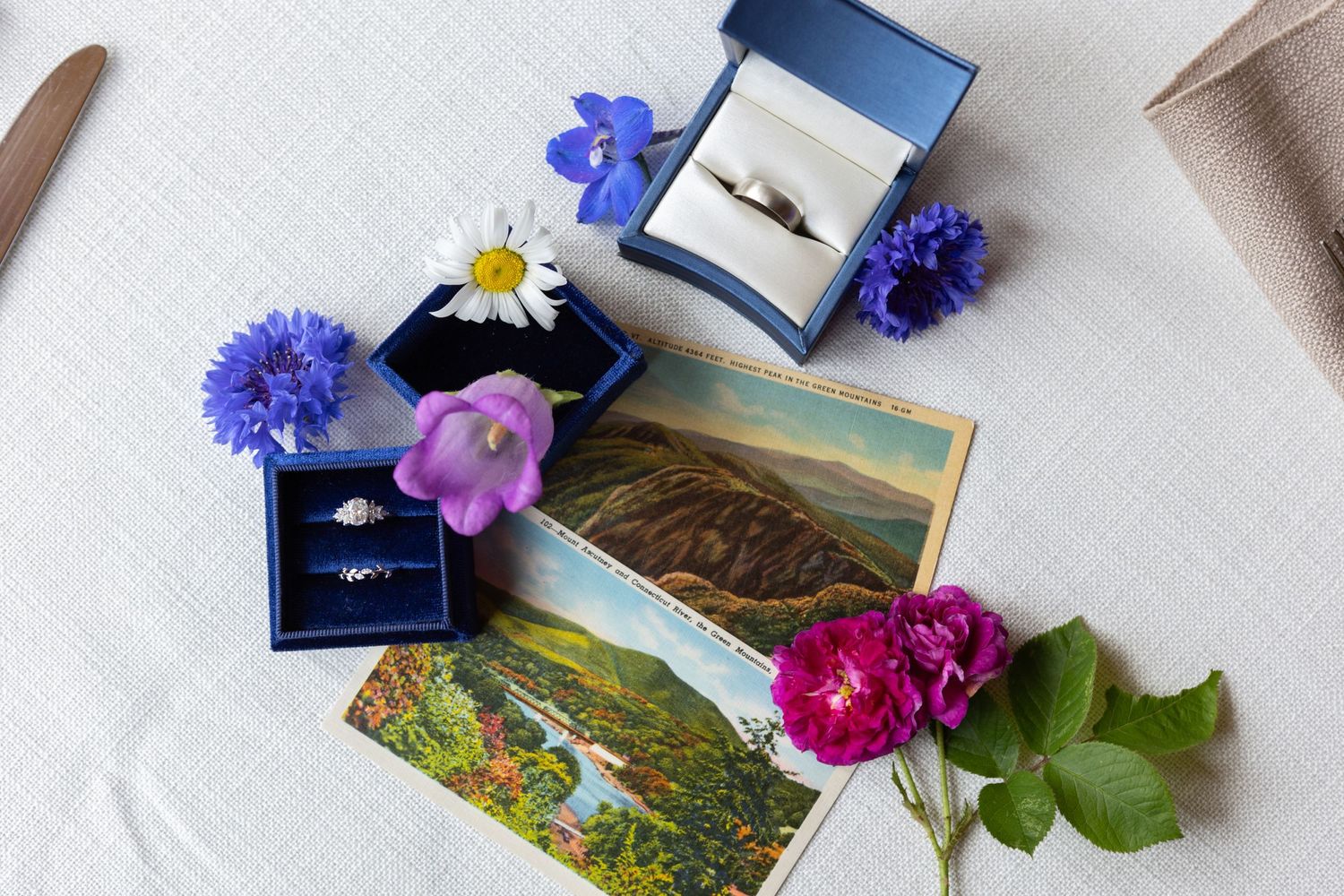 Wedding ring box with blue cornflowers, pink roses and vintage landscape postcards arranged on white fabric.