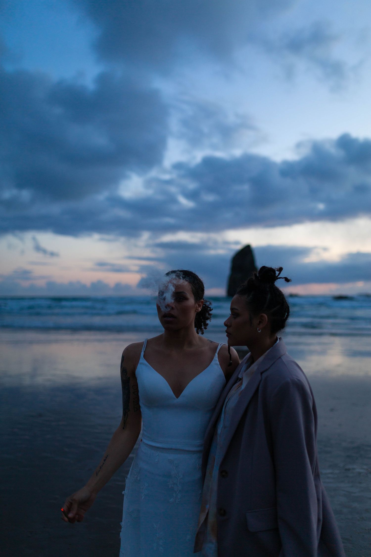 A couple stands together on a beach during blue hour with stormy clouds overhead.