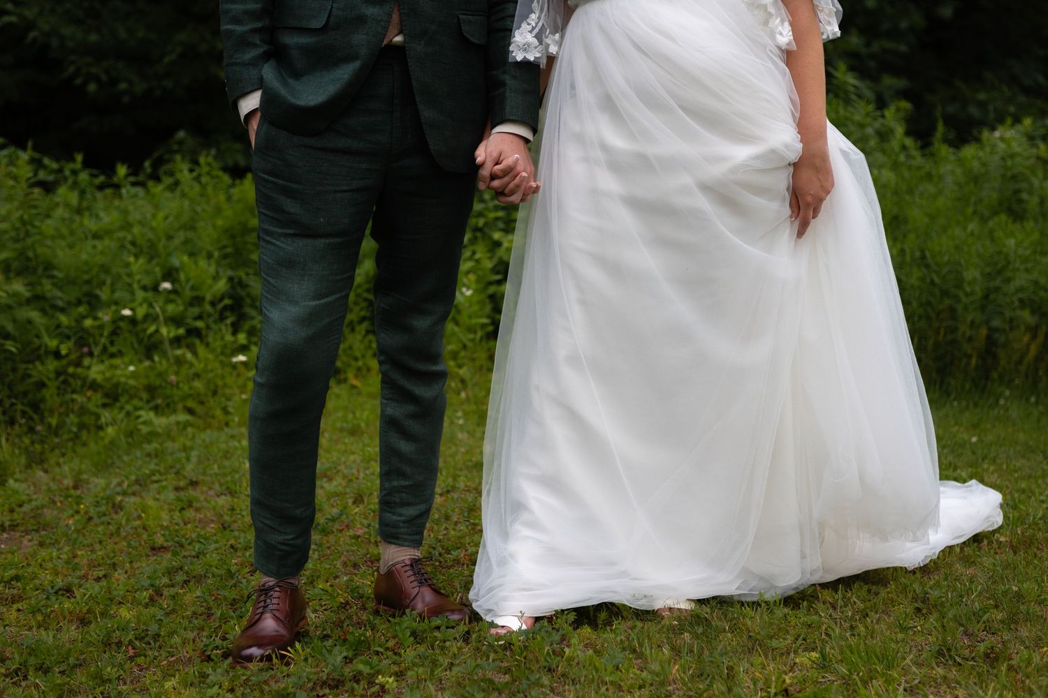 A wedding couple standing on grass, showing only their legs with the groom in a dark suit and bride in a white dress.