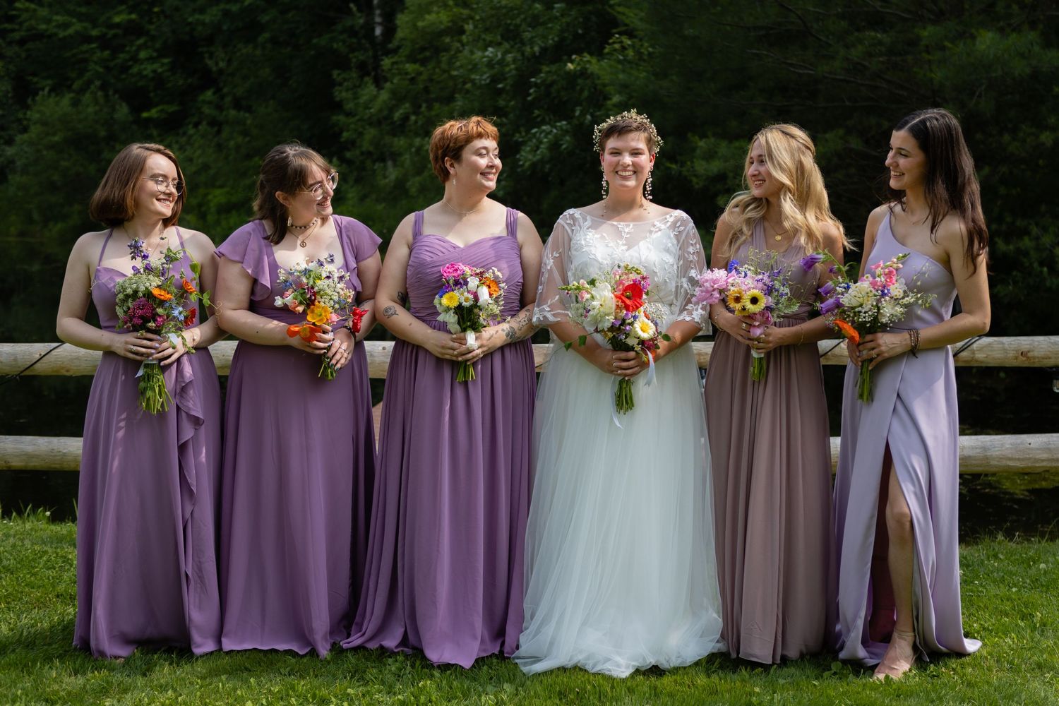 Bridesmaids in purple dresses pose with bride in white gown holding colorful bouquets outdoors.