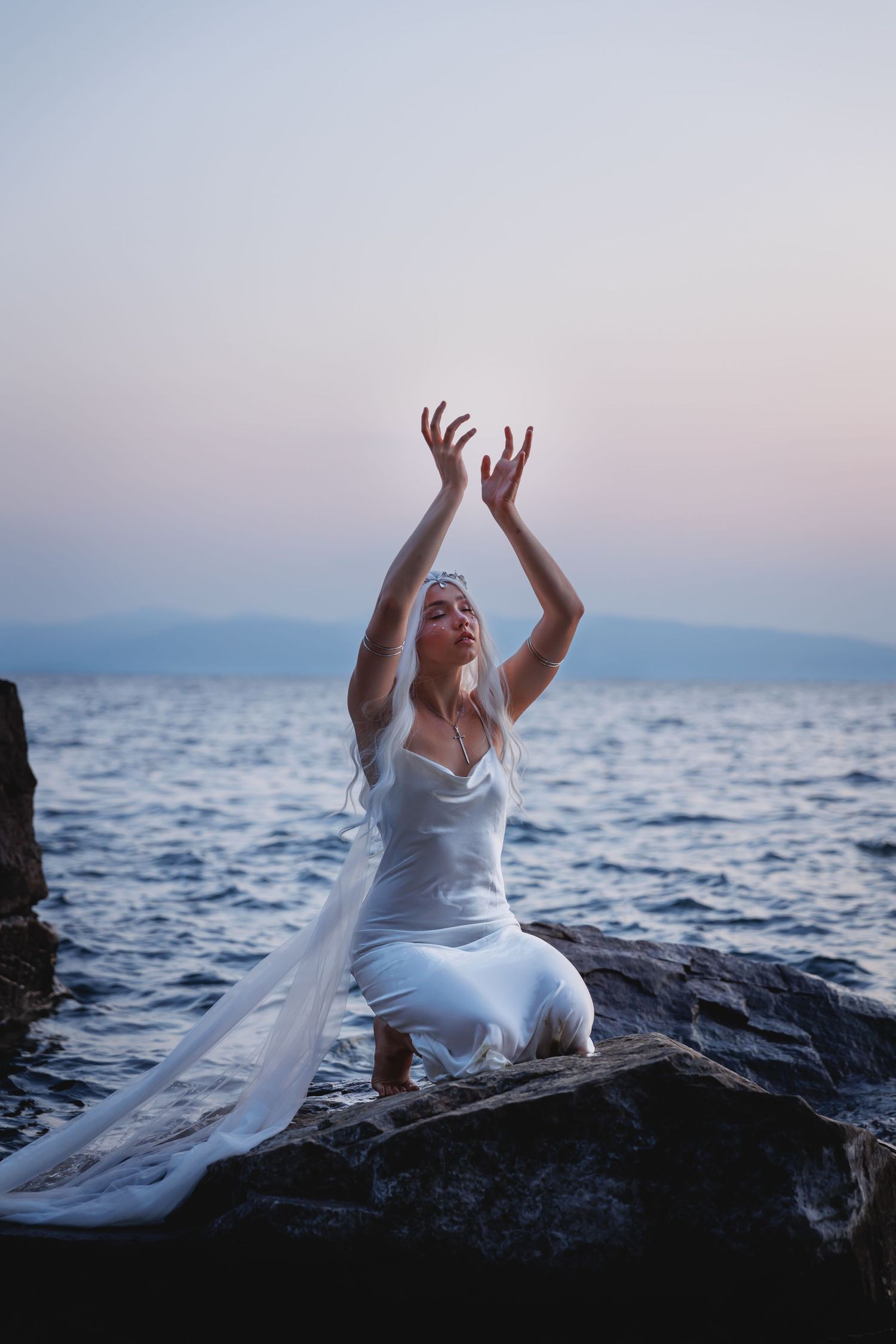 Blonde woman in white gown raises arms skyward while kneeling on rocks at twilight lake.