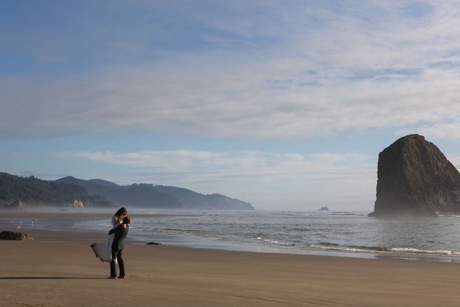 People walking along a misty Pacific Northwest beach with dramatic coastal cliffs and waves in the background.