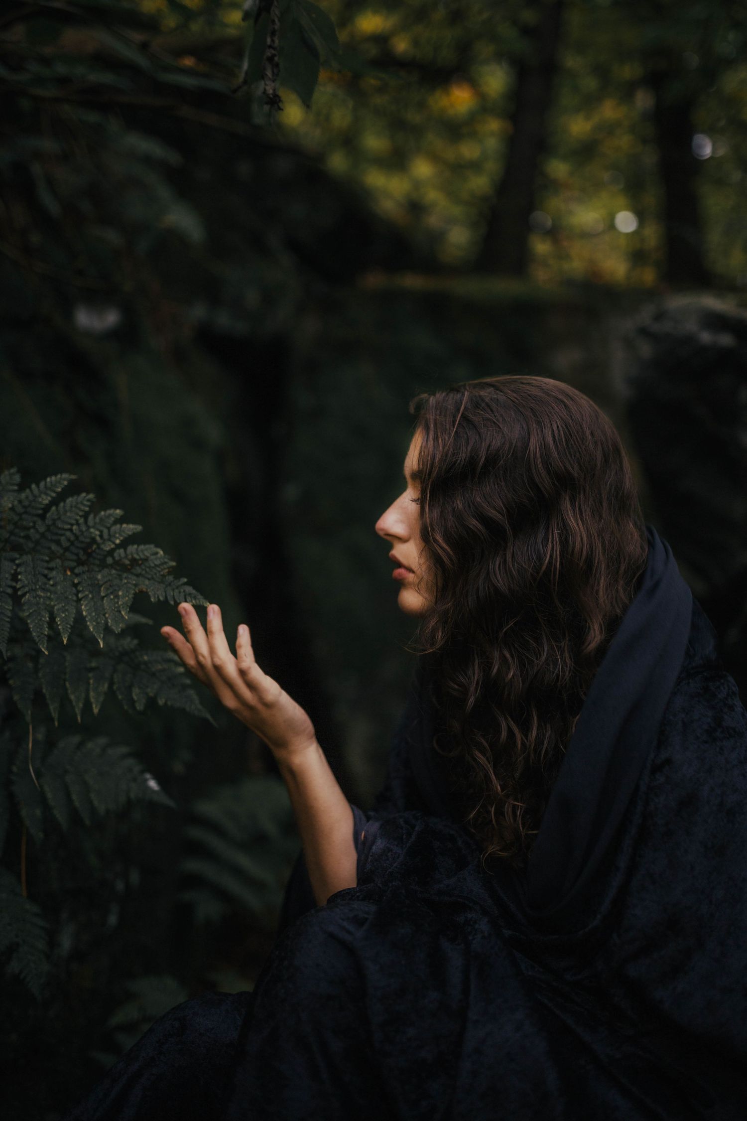 A dark-haired woman in a navy cloak gently touches a fern leaf in a moody forest setting.