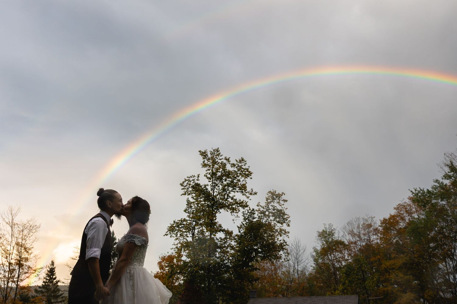 A couple kisses beneath a stunning full rainbow arching over autumn trees at golden hour.
