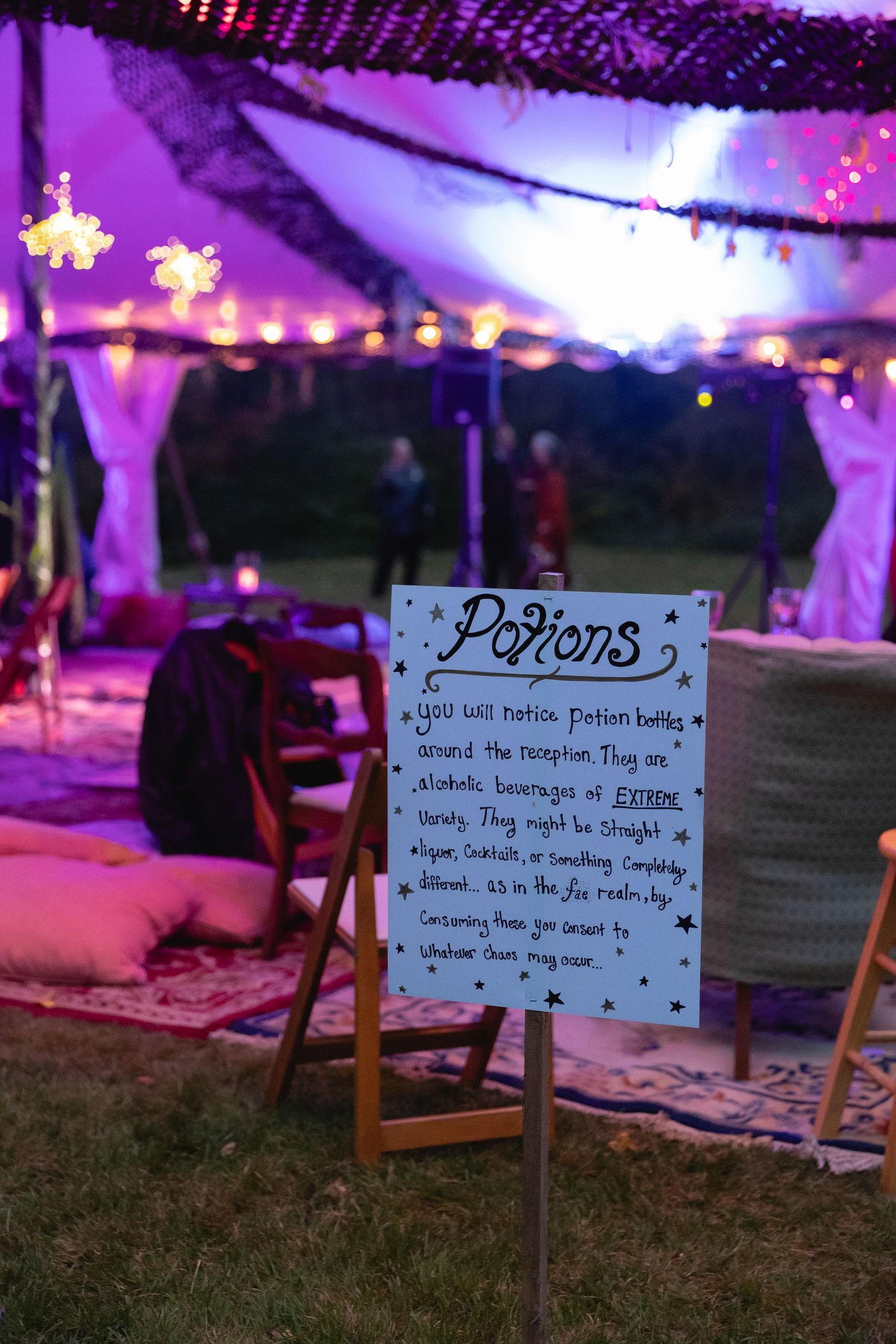 Whimsical wedding potions sign on easel inside purple-lit bohemian reception tent with floor cushions.