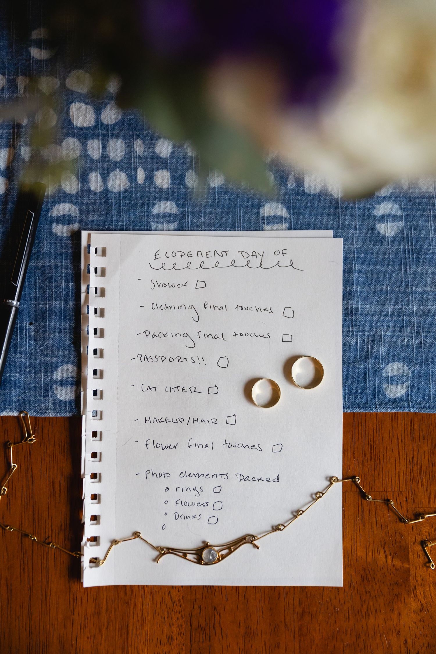 A handwritten wedding checklist on notepad paper with two gold wedding rings placed on it against a blue and wooden surface.