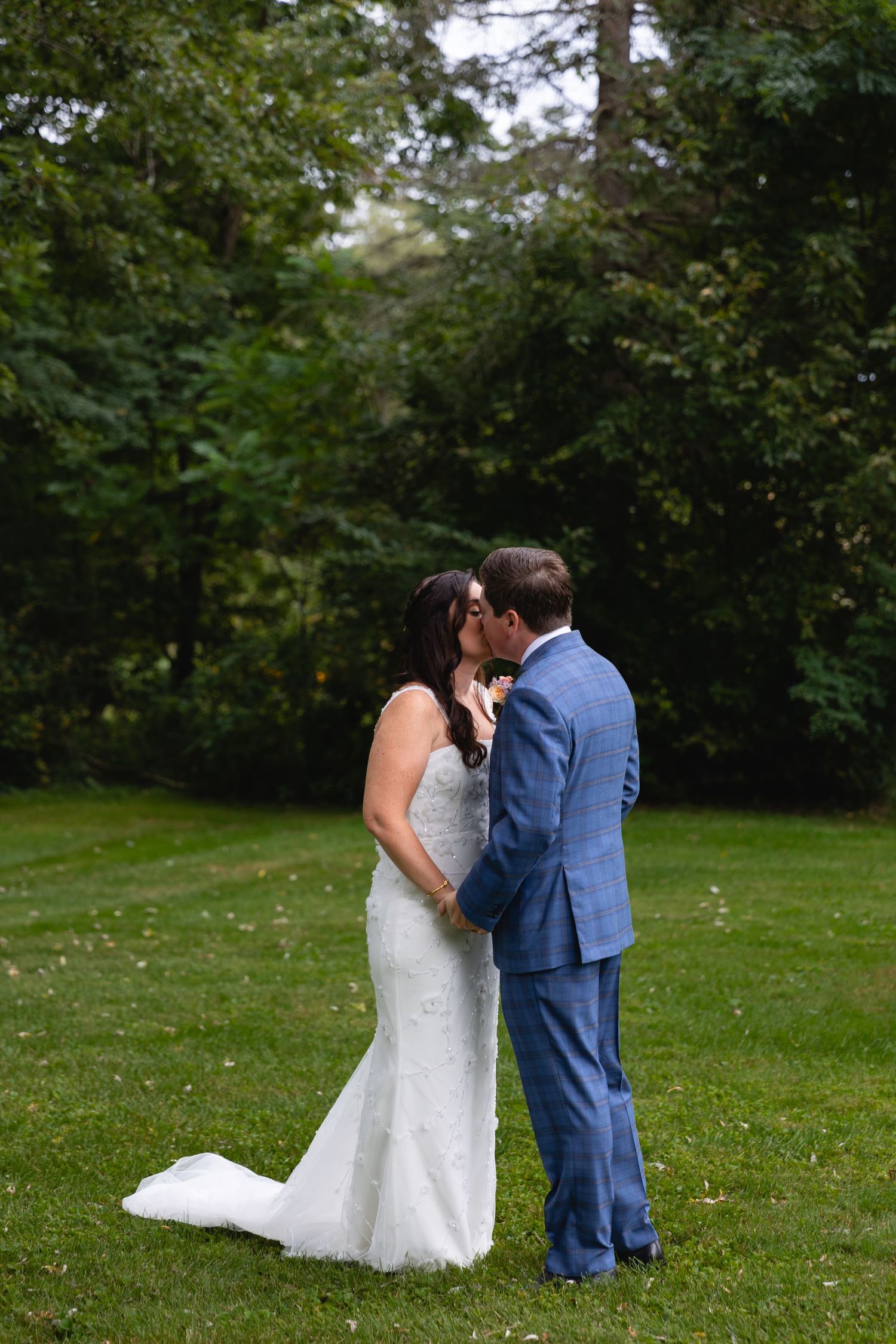 A romantic outdoor wedding photo showing a couple in white dress and blue suit embracing on a green lawn.