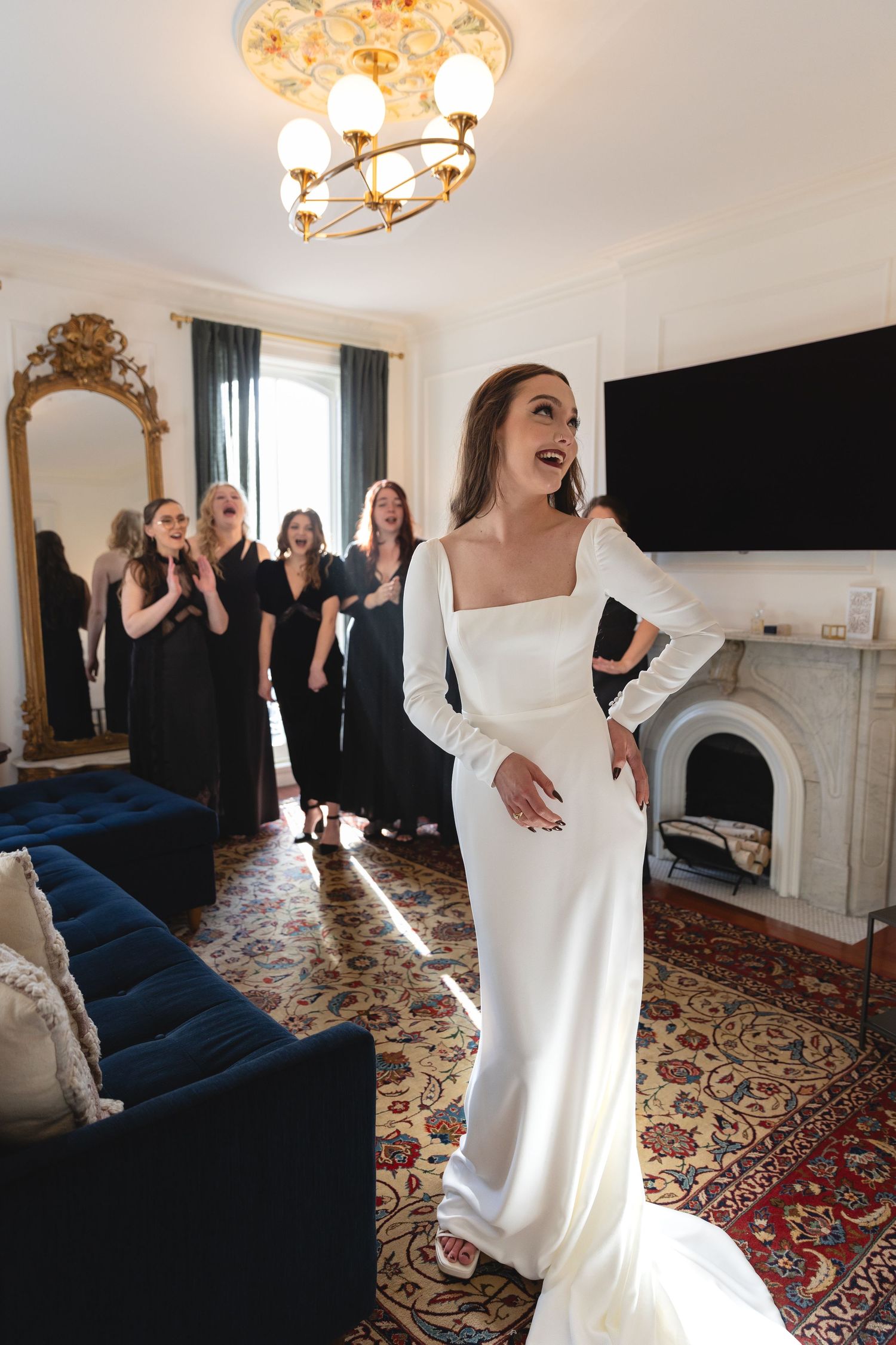 A person in a white gown stands in front of others wearing black dresses in an elegant room with a fireplace.
